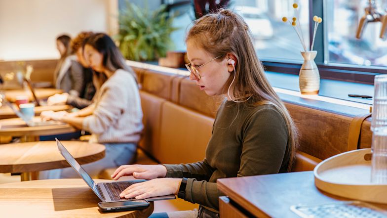 Person with earbuds working on laptop in cozy café with brown leather seating and natural light.