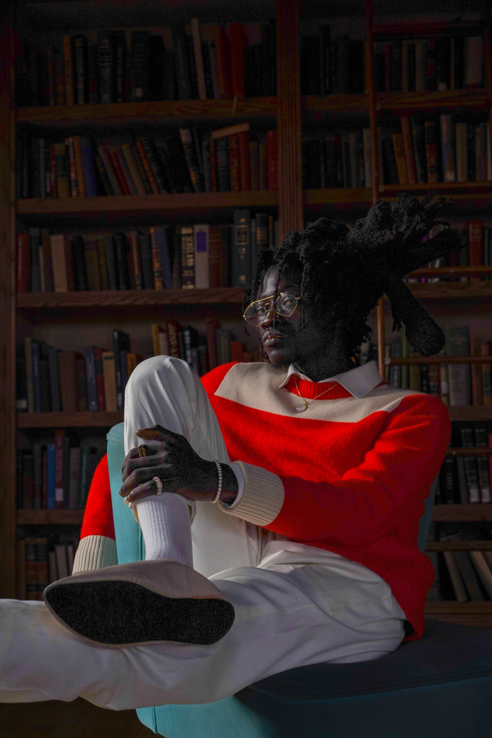 A model wearing a bright red sweater, white trousers and glasses sits on a chair in front of a bookshelf and looks at the camera, shot by Maggie Hinton with a Sony A9