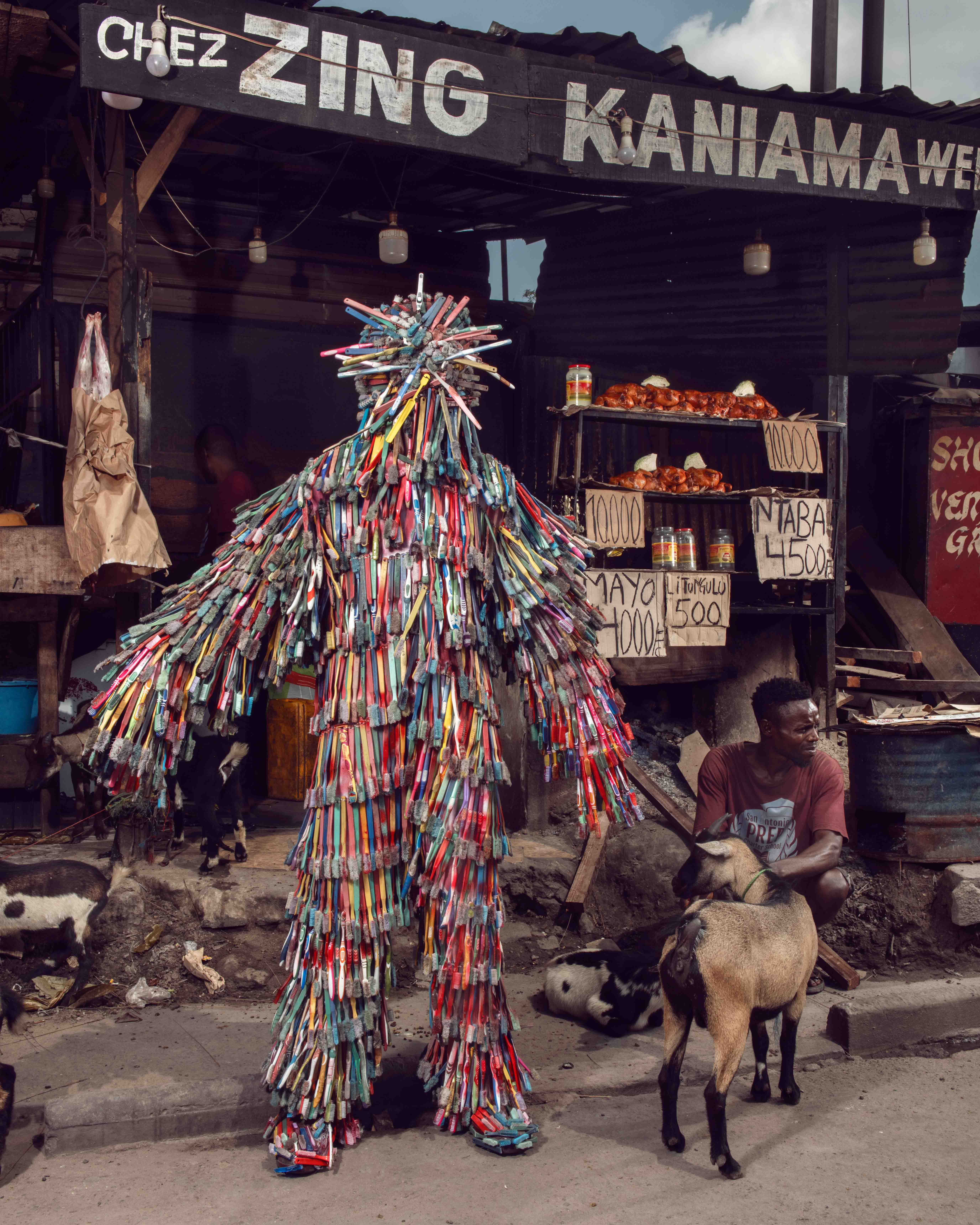 Eine schillernde Gestalt aus Stephan Gladieus Homo Detritus, die in Zahnbürsten gekleidet an einem Fleischstand in Kinshasa steht.
