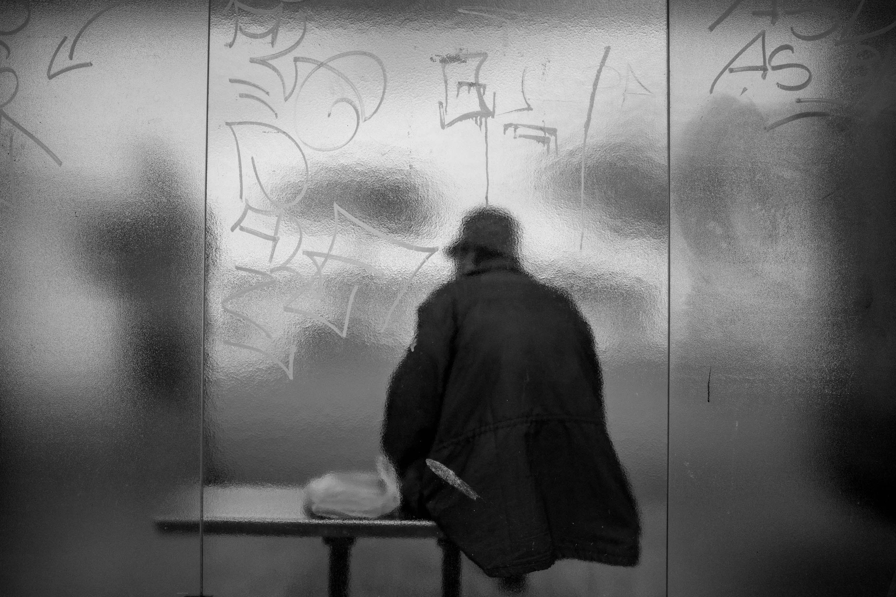 De figuur van een persoon zittend achter het gedeeltelijk doorschijnende glas van een bushalte bedekt met graffiti, zwart-wit straatfotografie door Ian Howorth