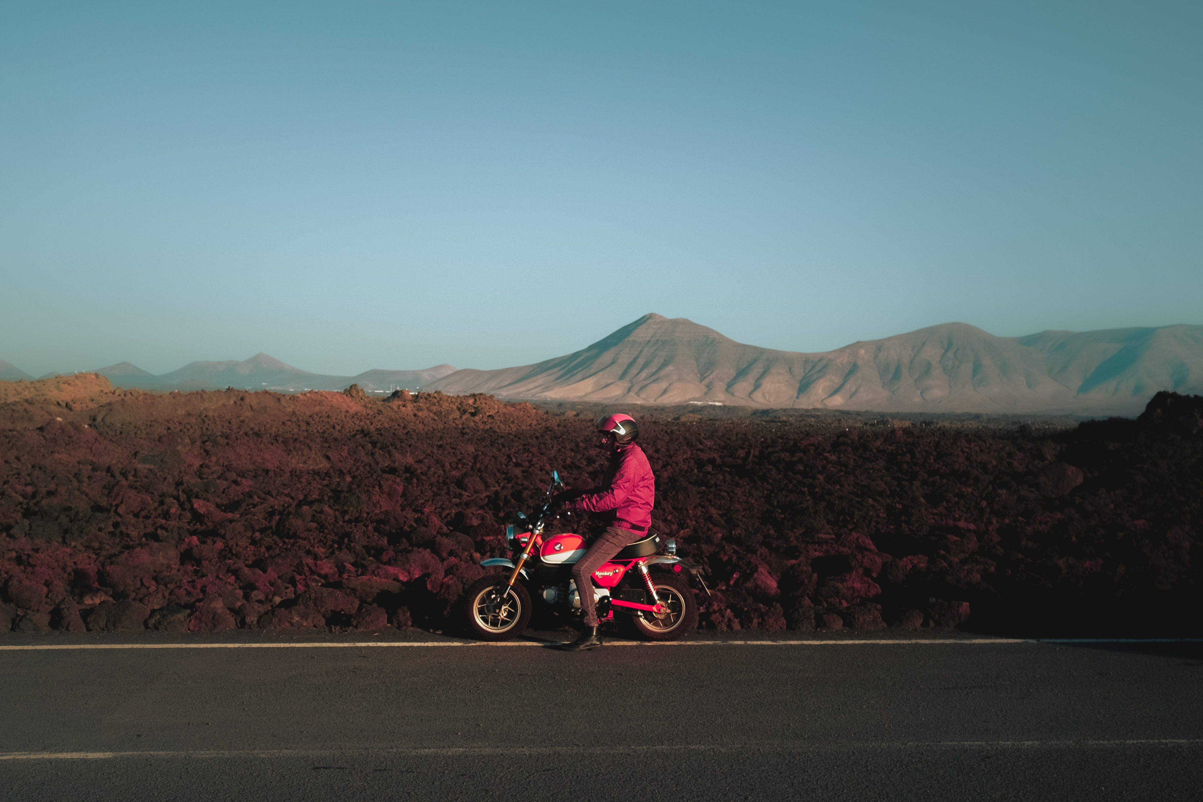 Infrarotfoto einer Person mit pinker Jacke auf einem pinken Motorrad auf einer von Bergen umgebenen Straße auf Lanzarote. Aufgenommen von Ian Howorth mit einer IR-konvertierten Fujifilm X-T1.