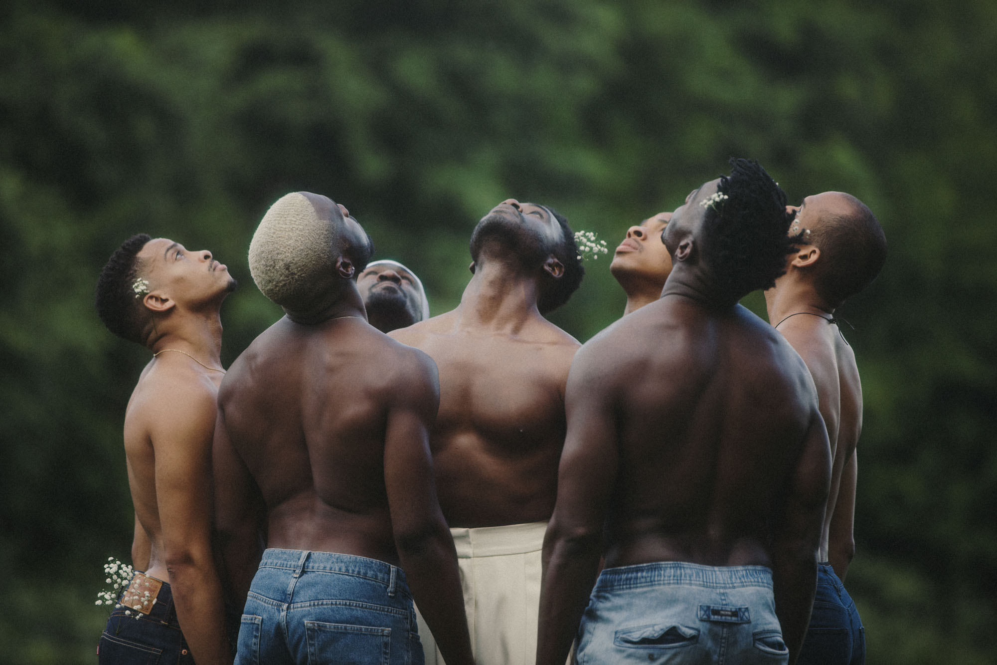 A group of models looking curiously up at the sky, some have white flowers in their hair, with out-of-focus trees in the background