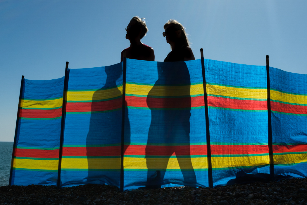 Two people standing behind a windbreaker in the harsh midday sun
