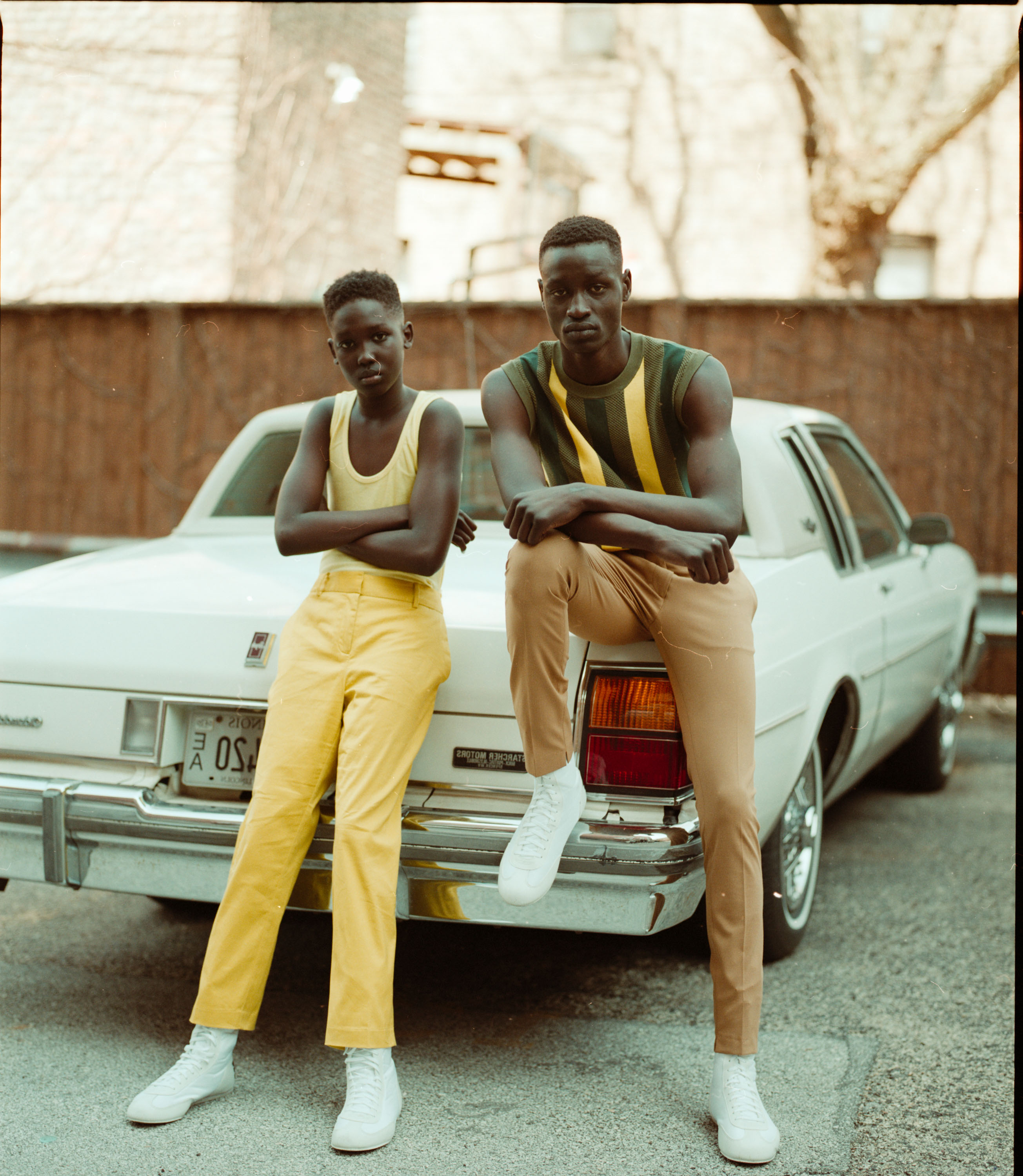 Two people pose for a portrait leaning against an old car