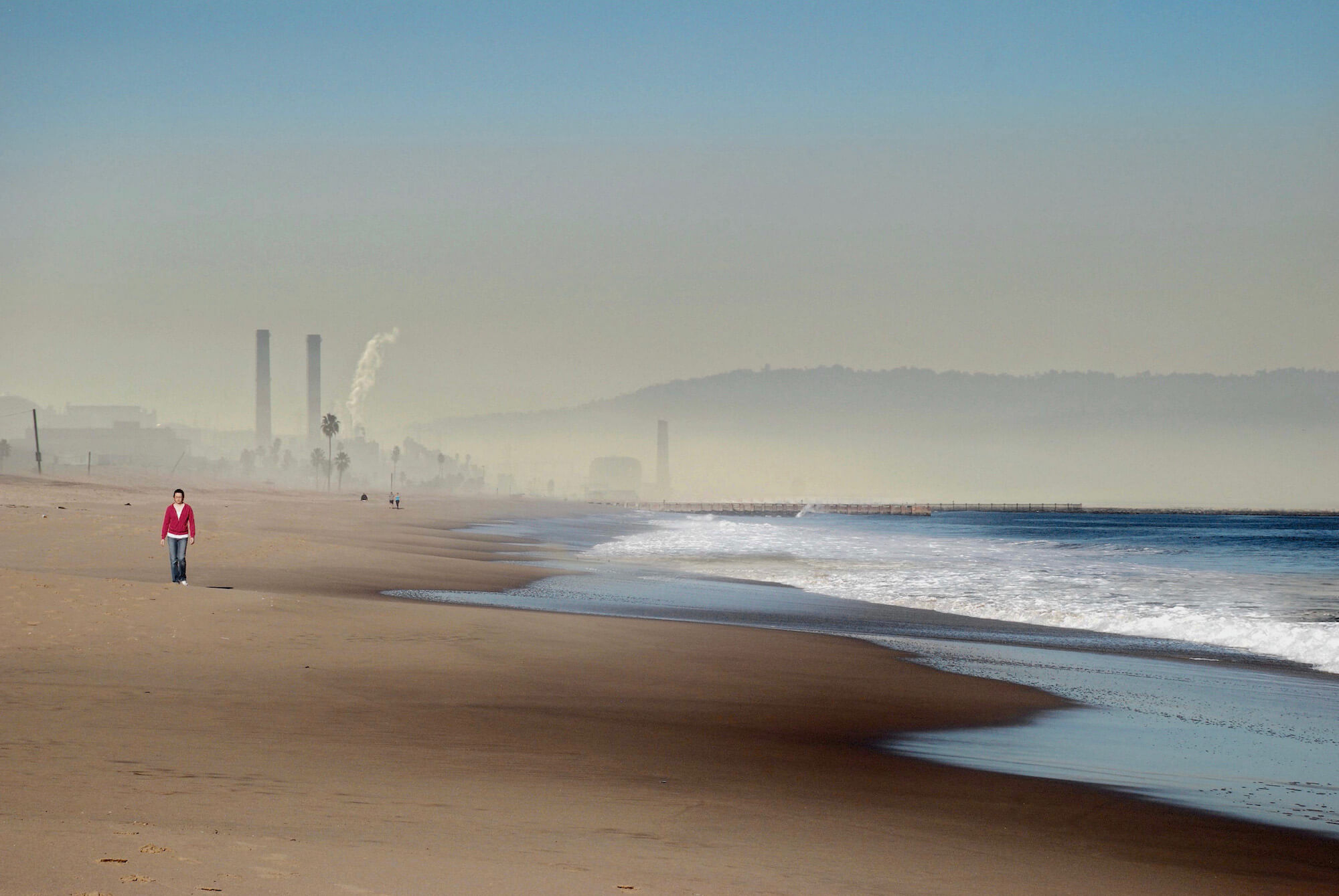 Man walking along the beach with cashing waves from the sea and industrial steelworks in the background.