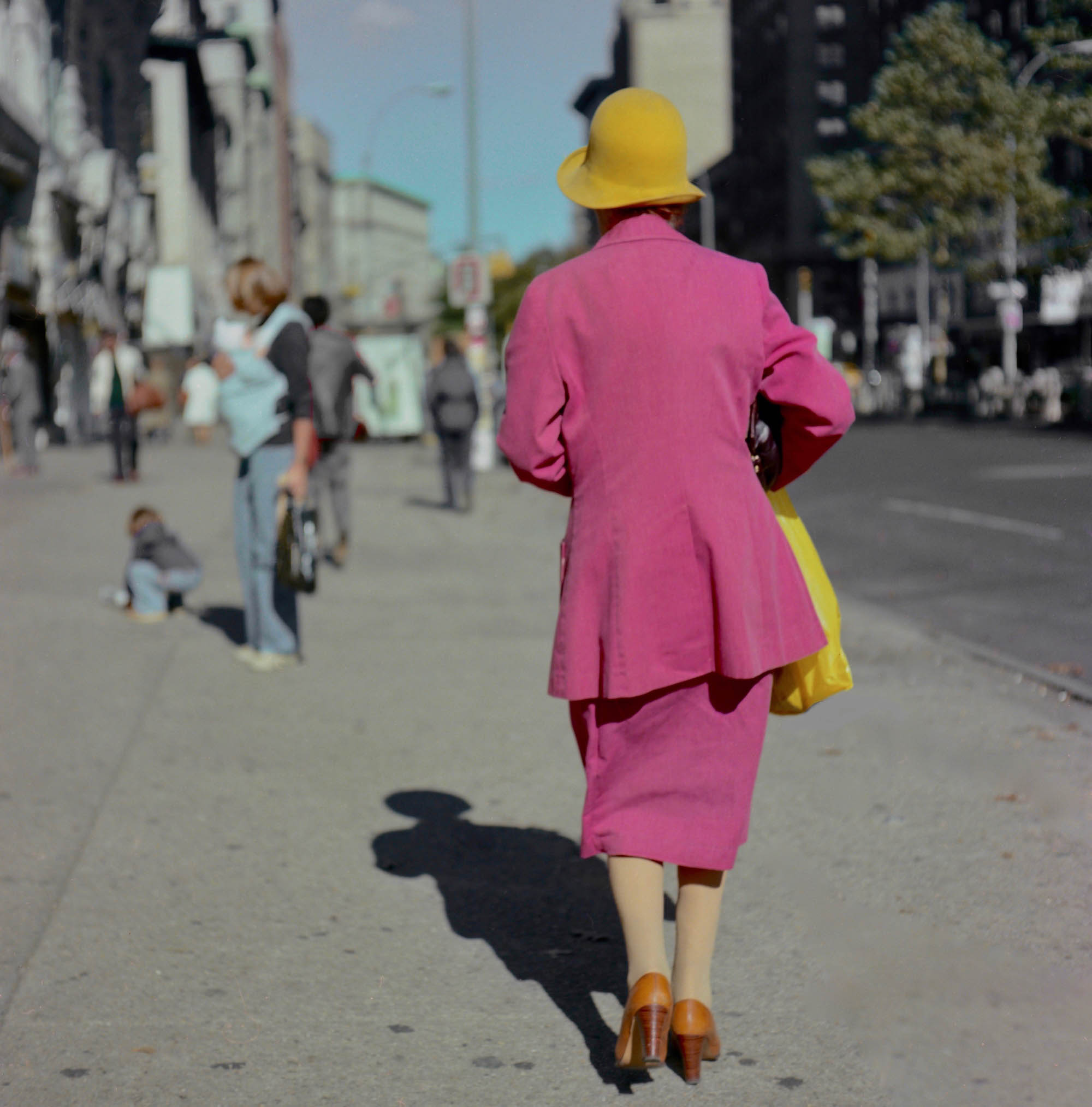 A lady wearing a pink cotton suit in the 70's walking away from the camera with a yellow hat.