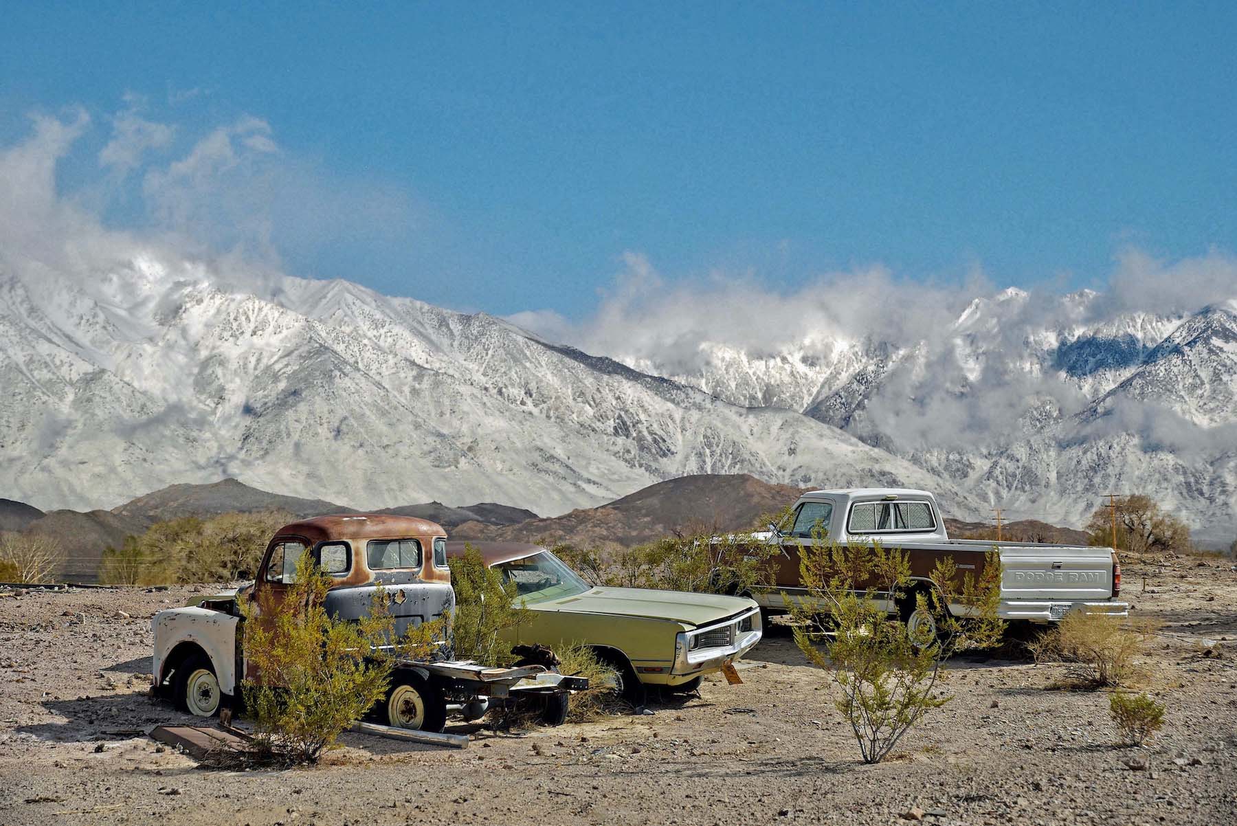 3 scrap pickup and two cars on a hill with mountains in the background.