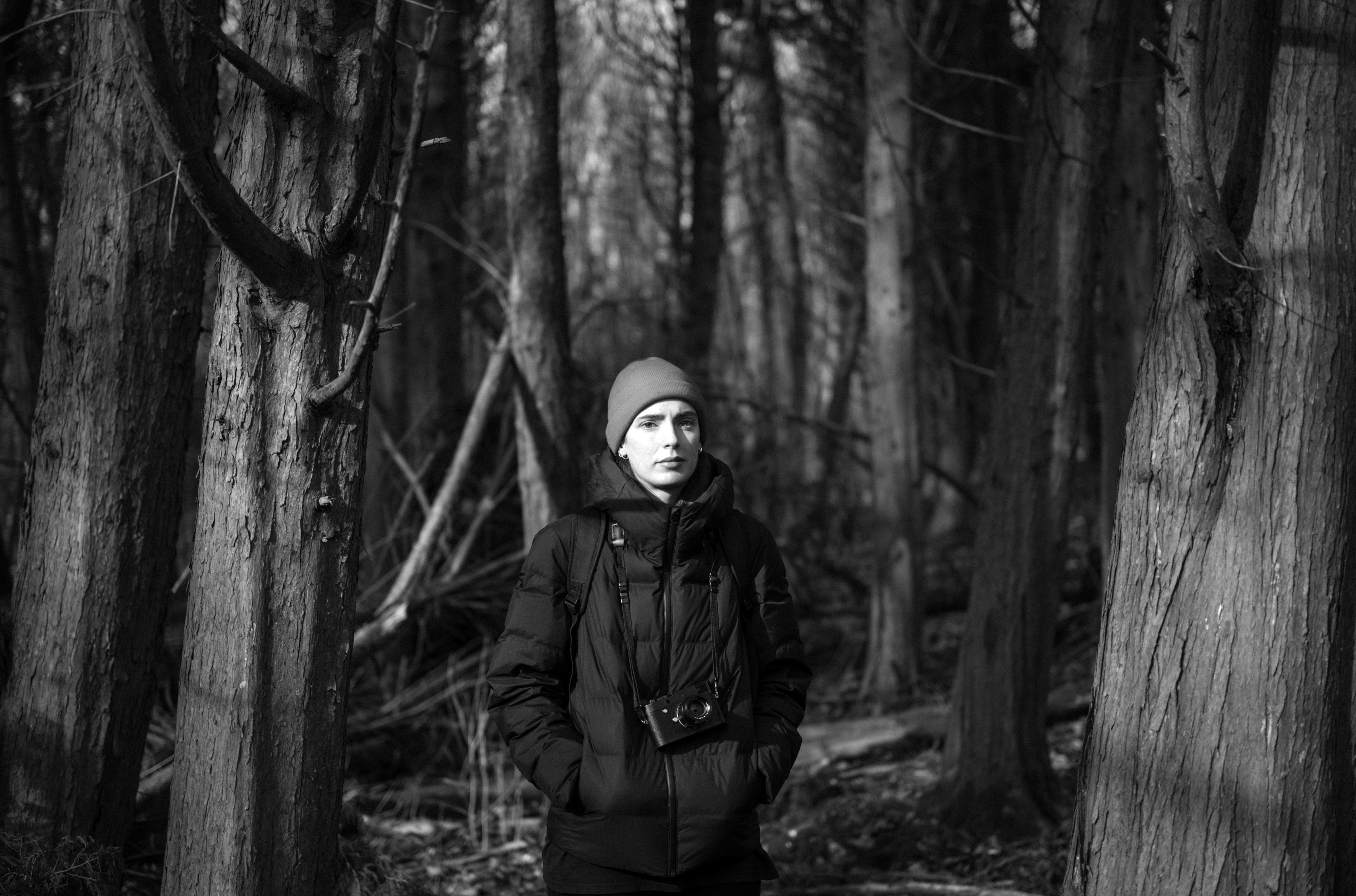 Foto in bianco e nero di un fotografo che indossa una giacca imbottita e un berretto, si trova davanti ad alcuni alberi e guarda verso la fotocamera, scattata con una Leica M Typ 246 Monochrom