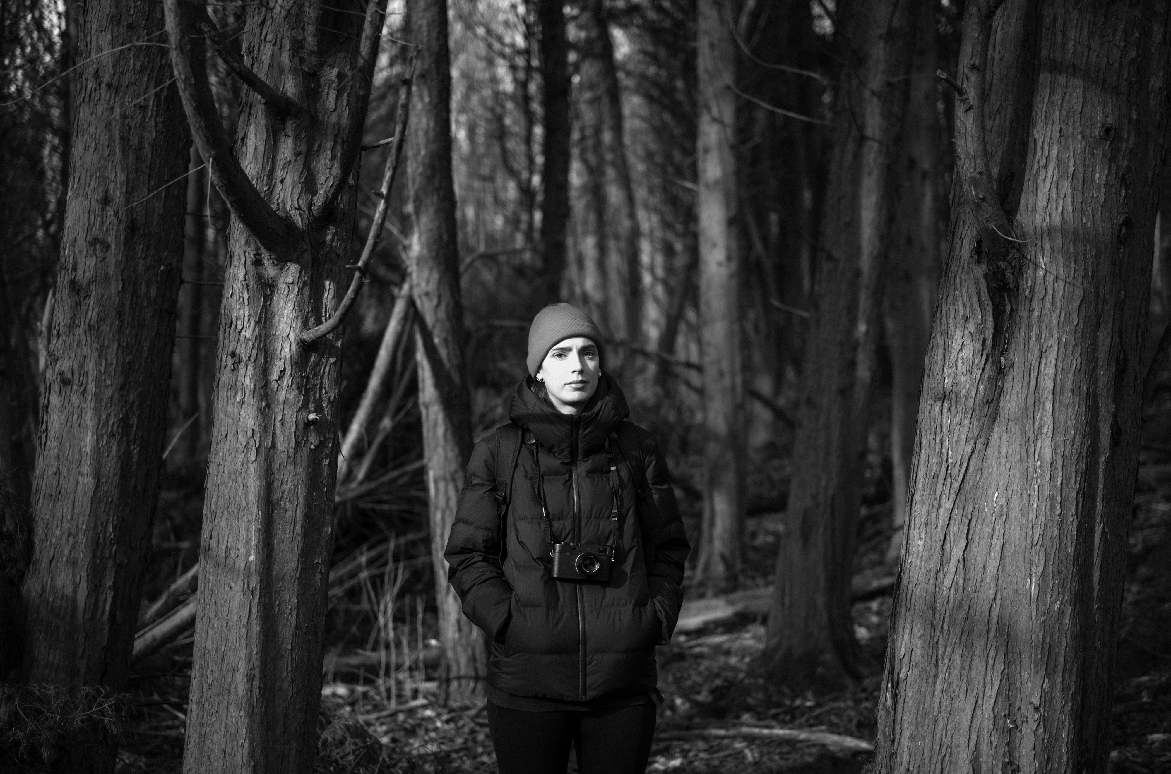 Foto in bianco e nero di un fotografo che indossa una giacca imbottita e un berretto, in piedi davanti ad alcuni alberi, che guarda verso la fotocamera, scattata con una Leica M10 Monochrom.