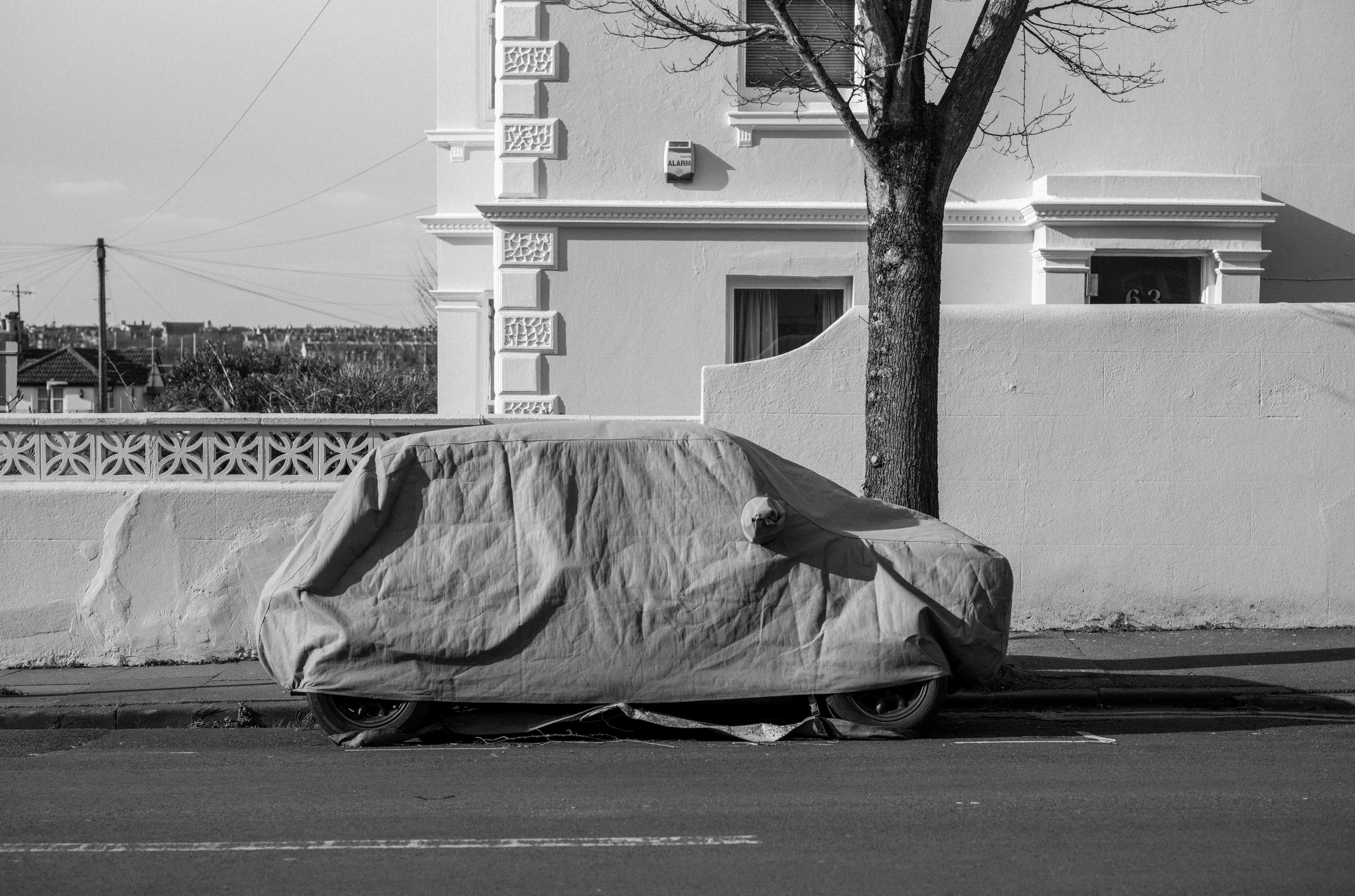 Ein Schwarz-Weiß-Foto von einem Auto, das mit einer Plane abgedeckt auf einer Straße neben einem Haus und einem Baum geparkt ist, aufgenommen mit einer Leica M Monochrom Typ 246