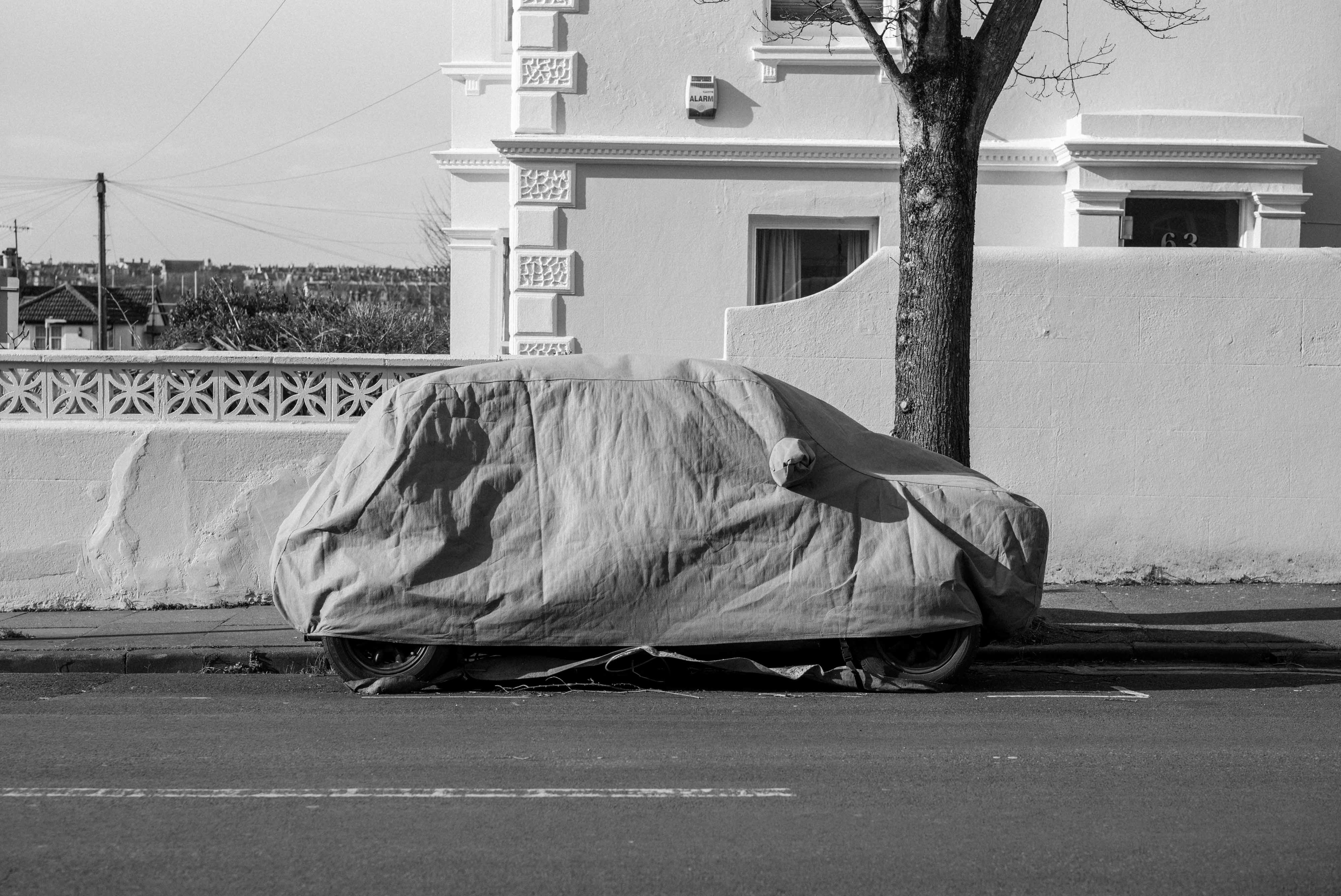 Ein Schwarz-Weiß-Foto von einem Auto, das mit einer Plane abgedeckt auf einer Straße neben einem Haus und einem Baum geparkt ist, aufgenommen mit einer Leica M10 Monochrom
