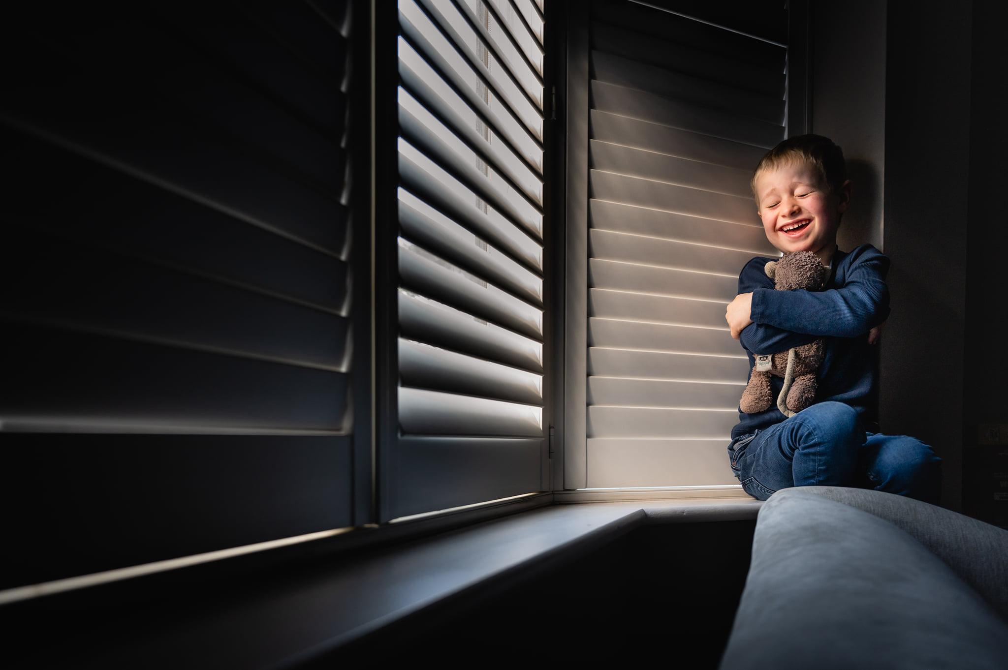A young child holding a teddy bear smiling while sat next to a window