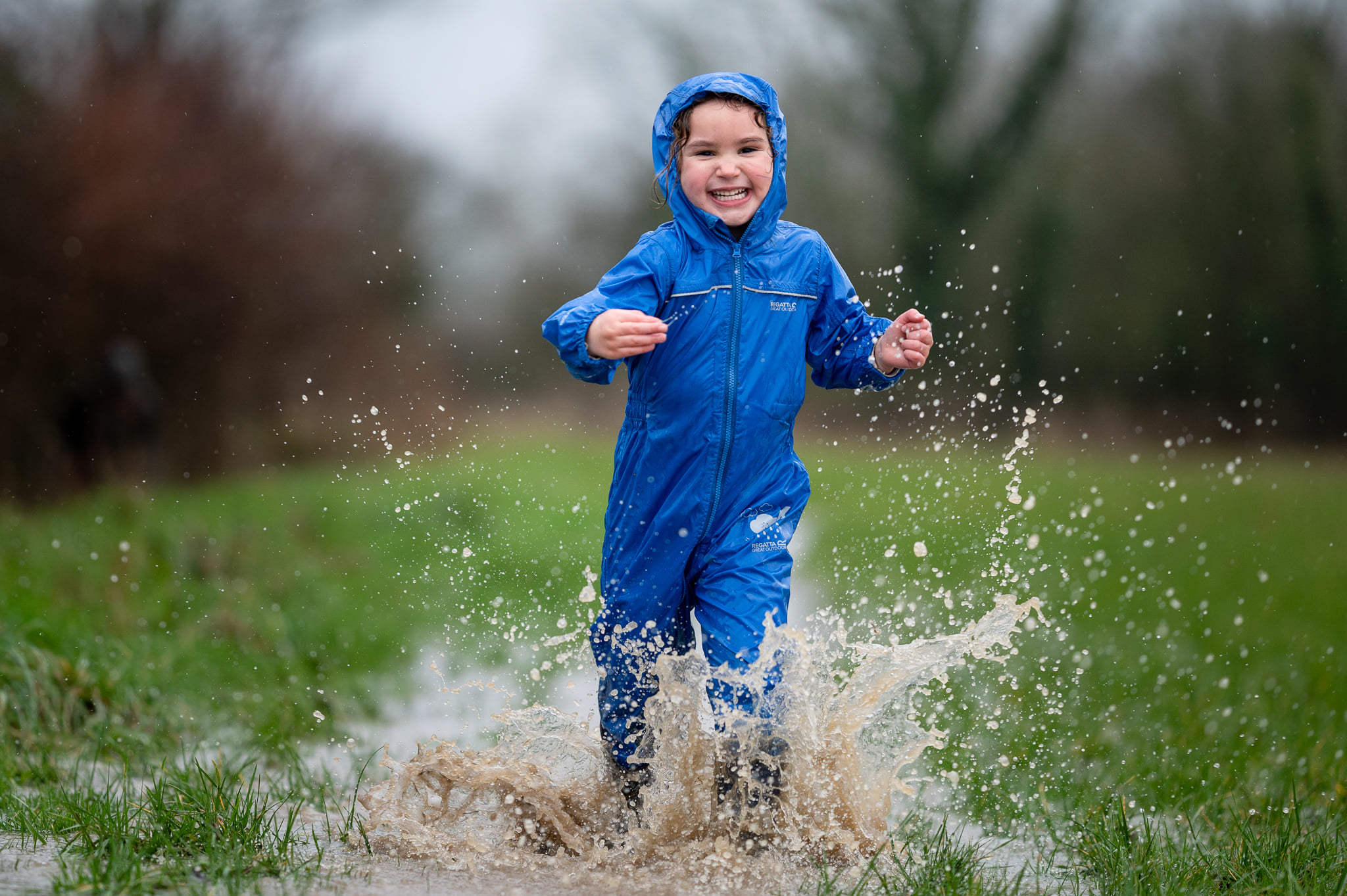 A kid in a blue waterproof onesie splashing through a puddle