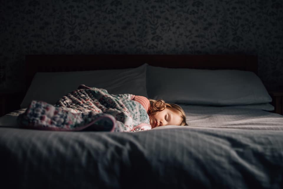 A young child sleeping on a bed, covered by a blanket, partially illuminated by a beam of light