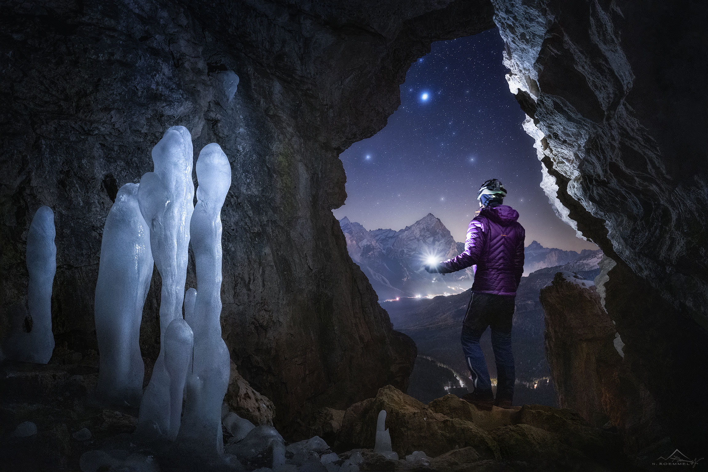 Blick aus einer Höhle mit Eisstrukturen. Eine stehende Person mit einer Taschenlampe blickt auf eine bergige Landschaft und den hellen Sternenhimmel.