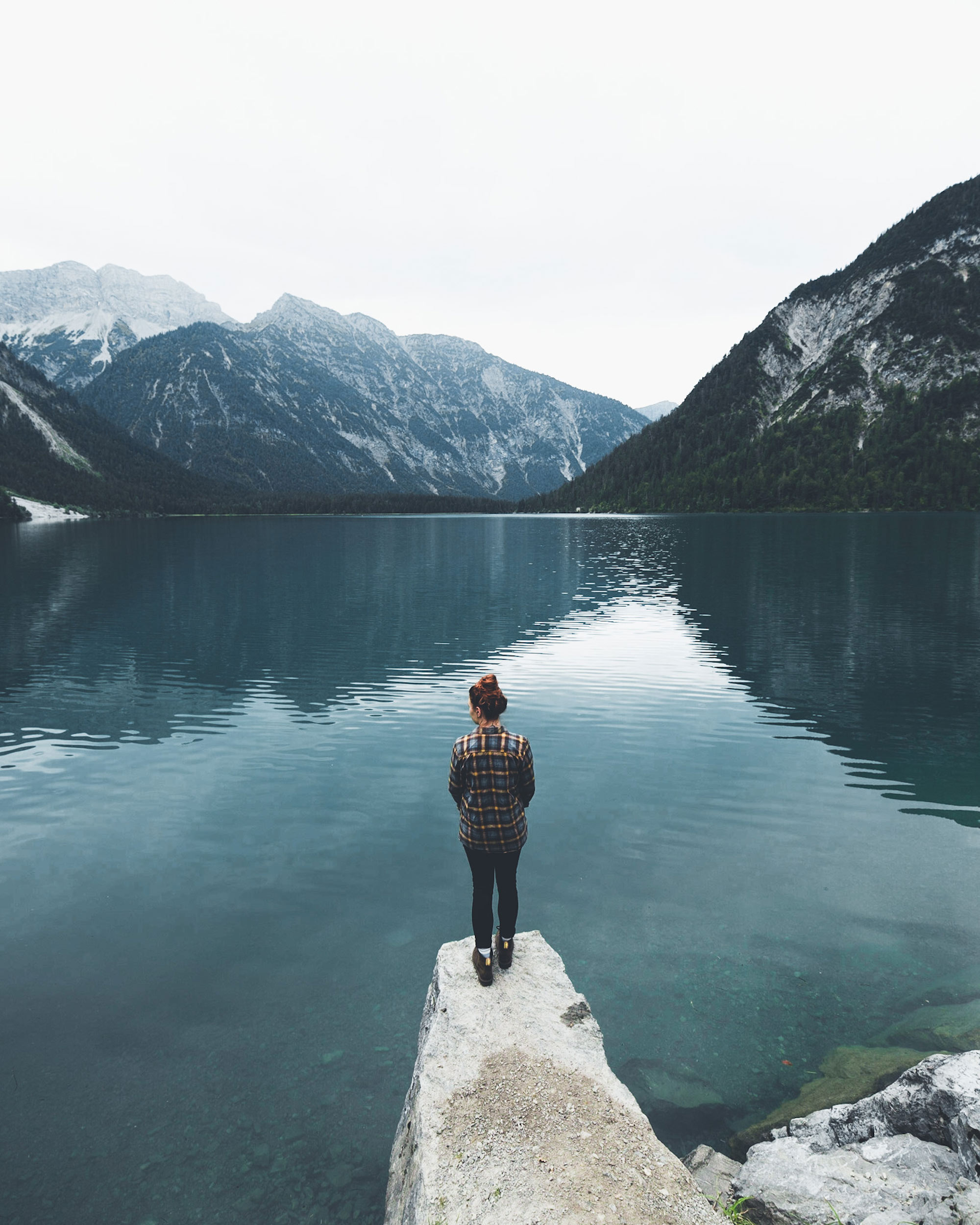 Person stands at the end of a cliff looking out over a mountain lake