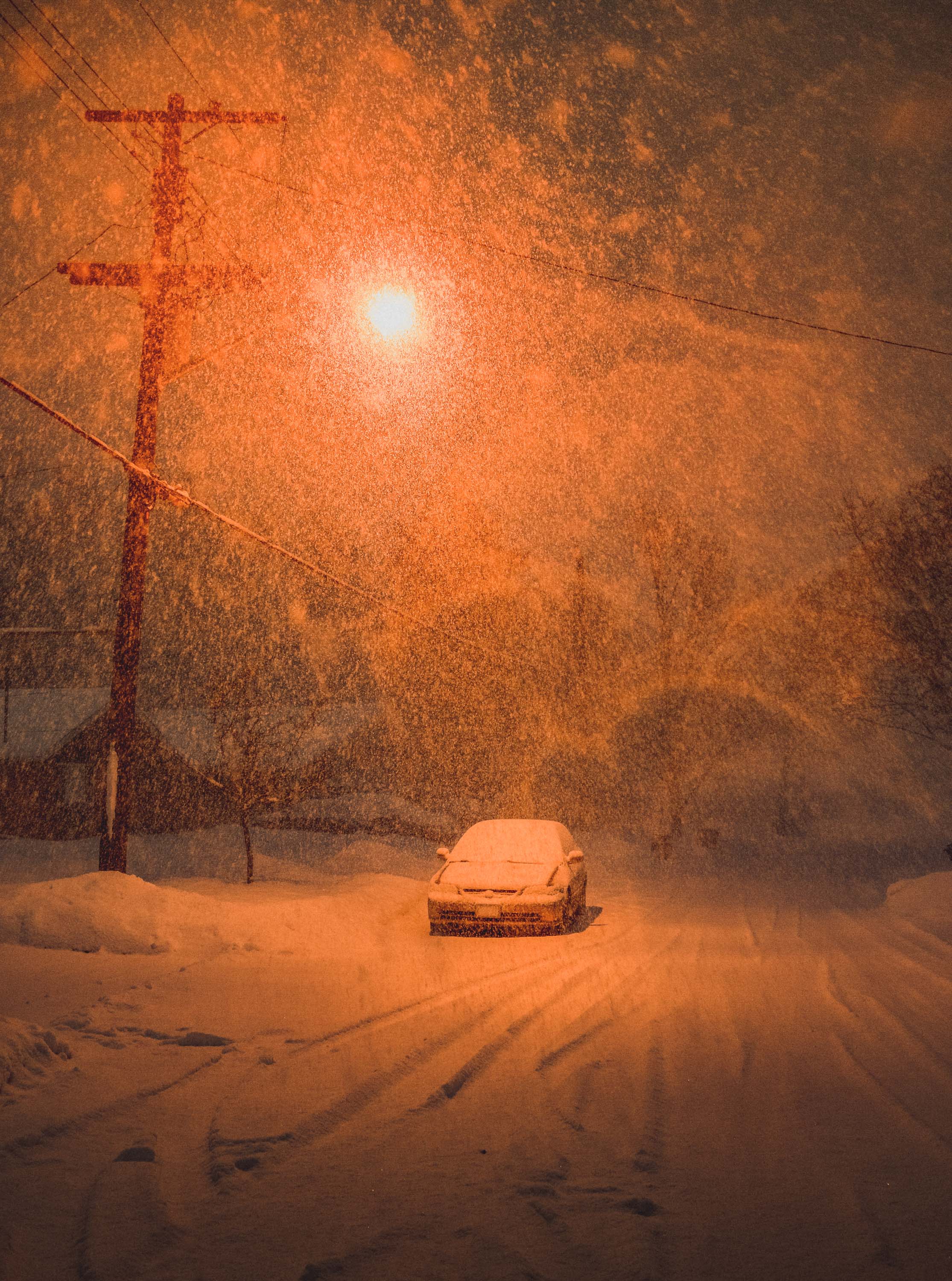 Heavily snow covered street with lonely car in the middle lit by a bright orange street lamp