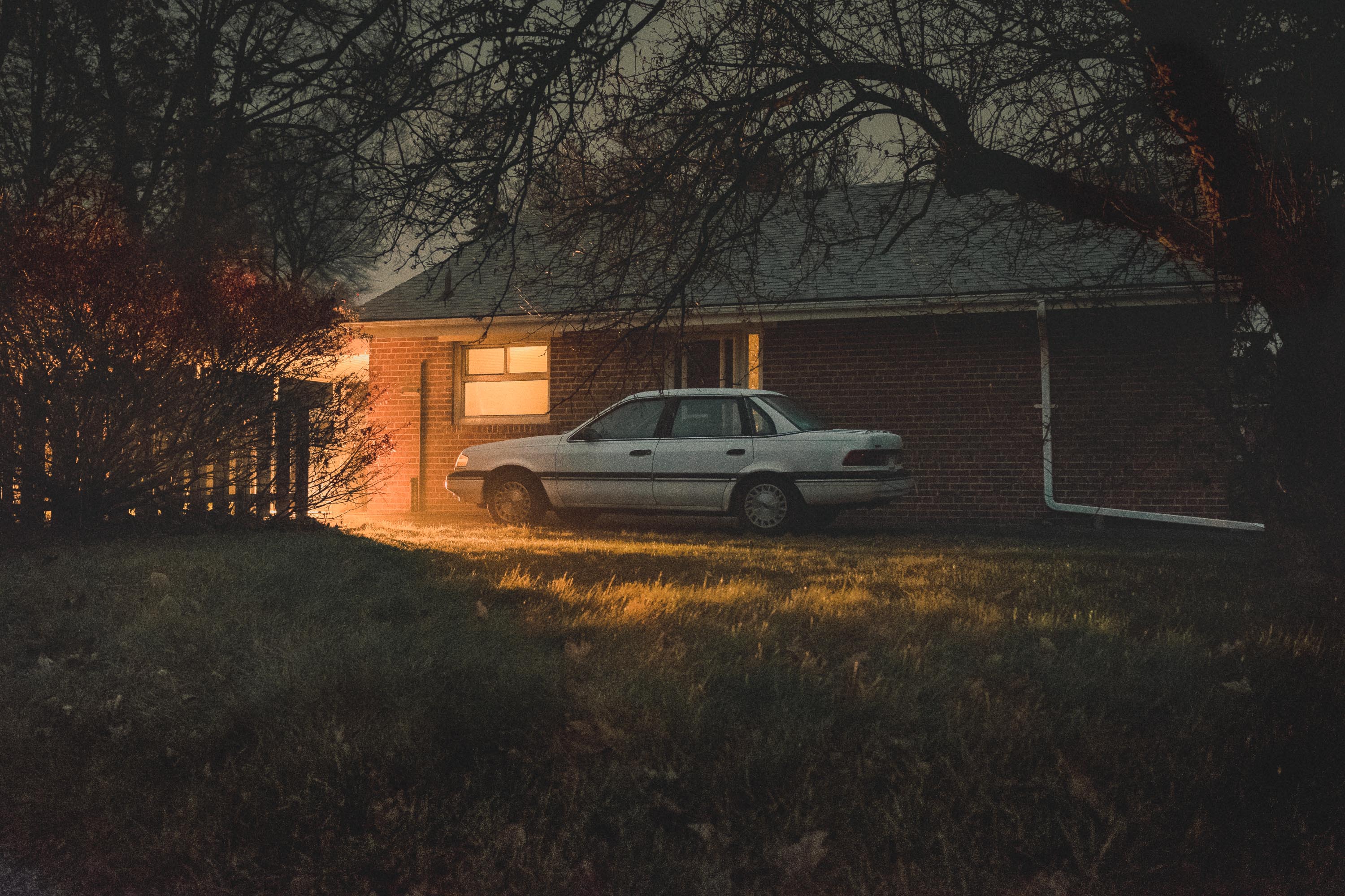 Car parked by a house with warm lights inside shining onto grass and the car