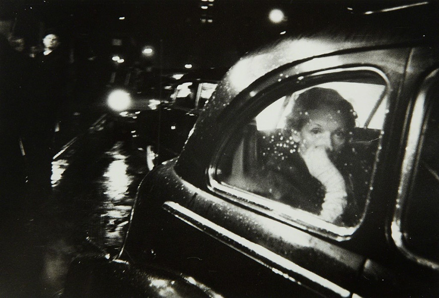 Black and white image of a woman looking through the window of a car while it rains outside