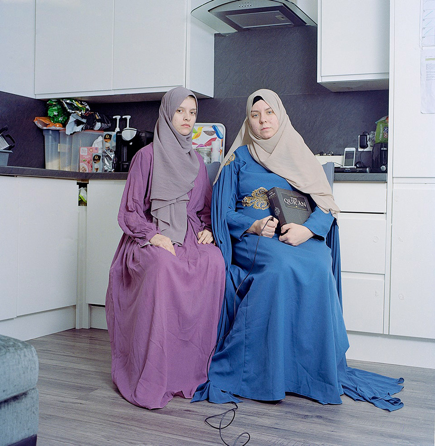 Two women wearing hijabs sitting down in a kitchen. Jodie Bateman @jodiembateman.