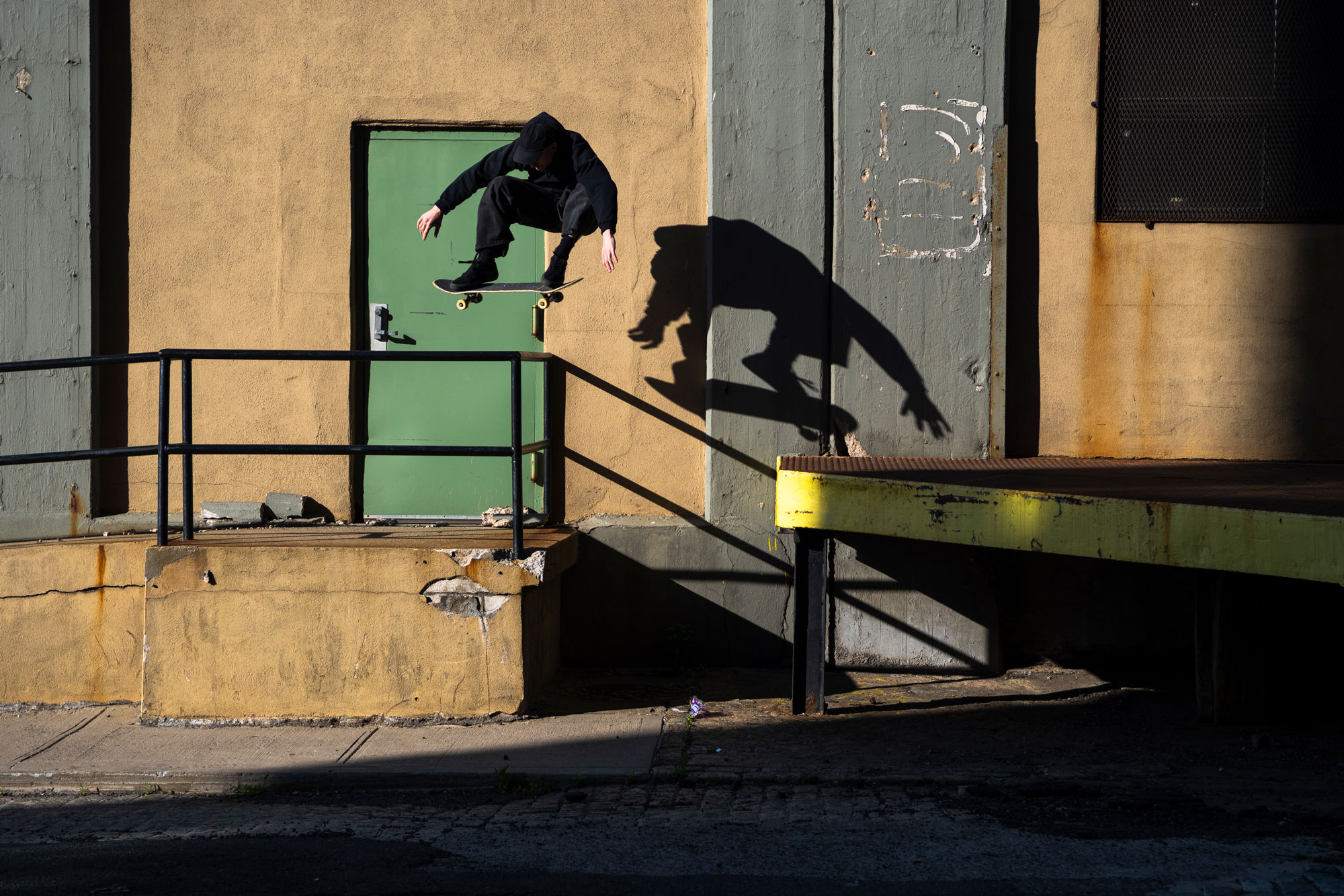 Skateboarder jumping over rail, with shadow on the wall. Shot by Jon Mecca.