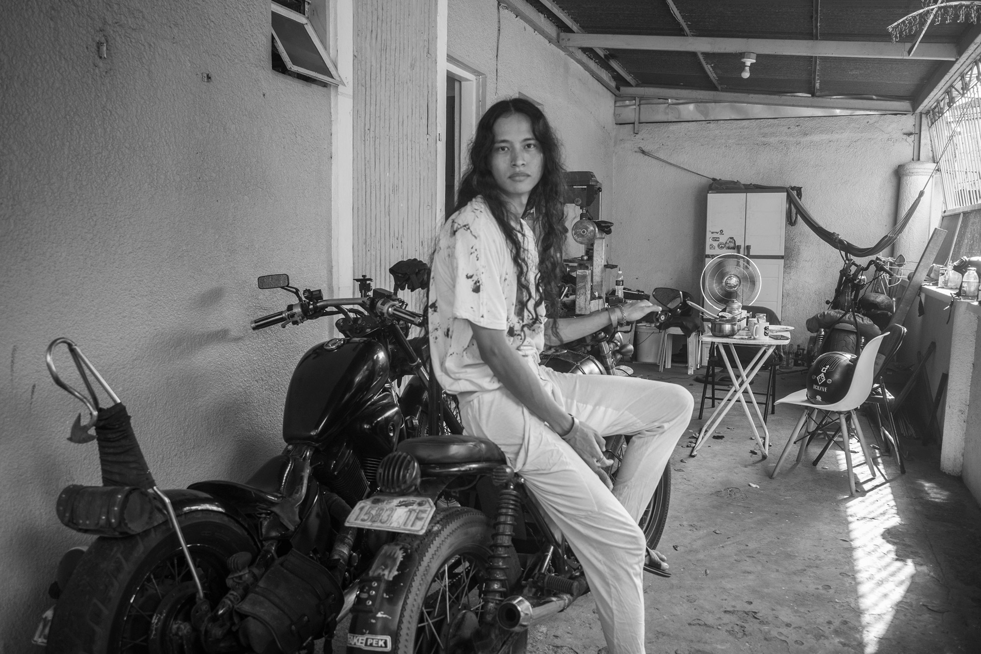 Black and white image of a person sitting on a motorbike in a garage. Shot by Regine David. 