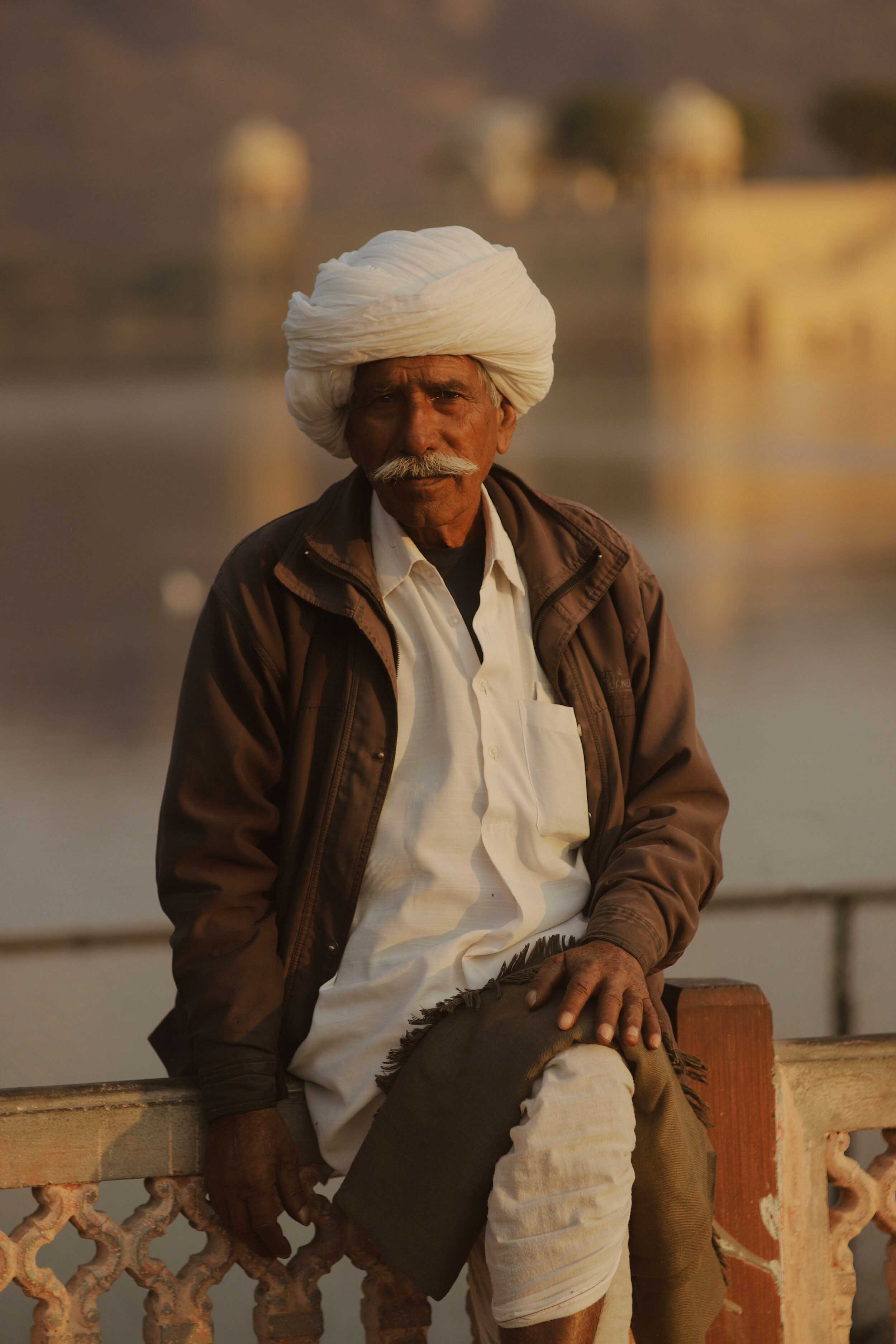 A person with a mustache and traditional headwear looks at the camera while sitting on a bridge with a river and a building in the background