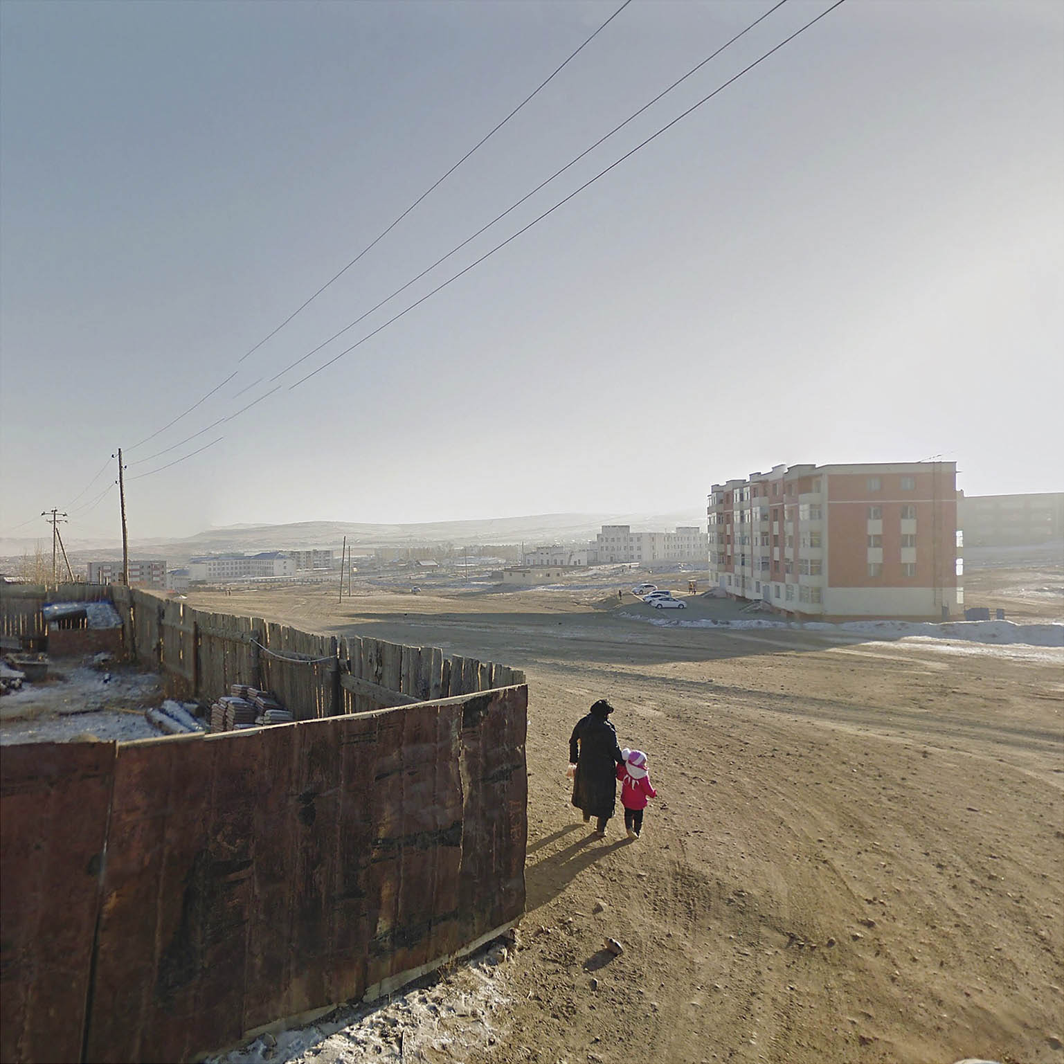 A parent and child walk by a wooden fence towards a building surrounded by sand.