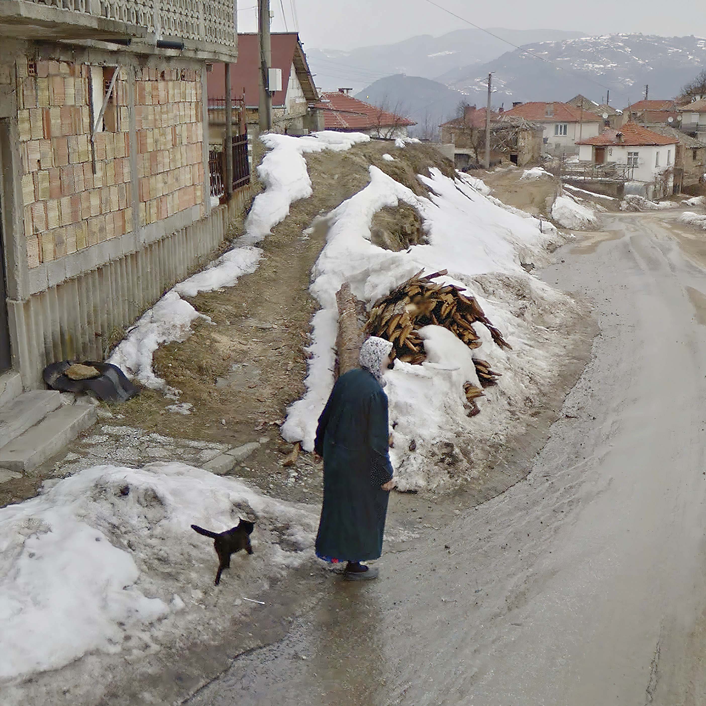 An elderly woman wearing a long turquoise coat and a head covering walks her cat along a slushy snowy road in a village surrounded by mountains  