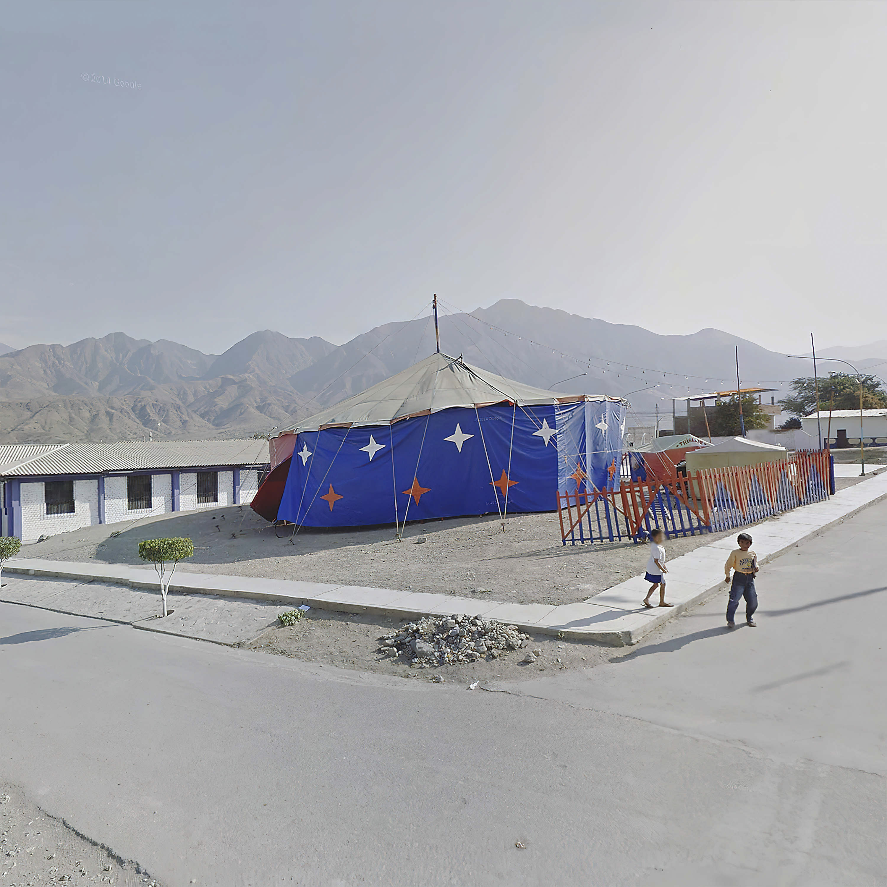Two children walk by a circus tent with a range of mountains in the background  