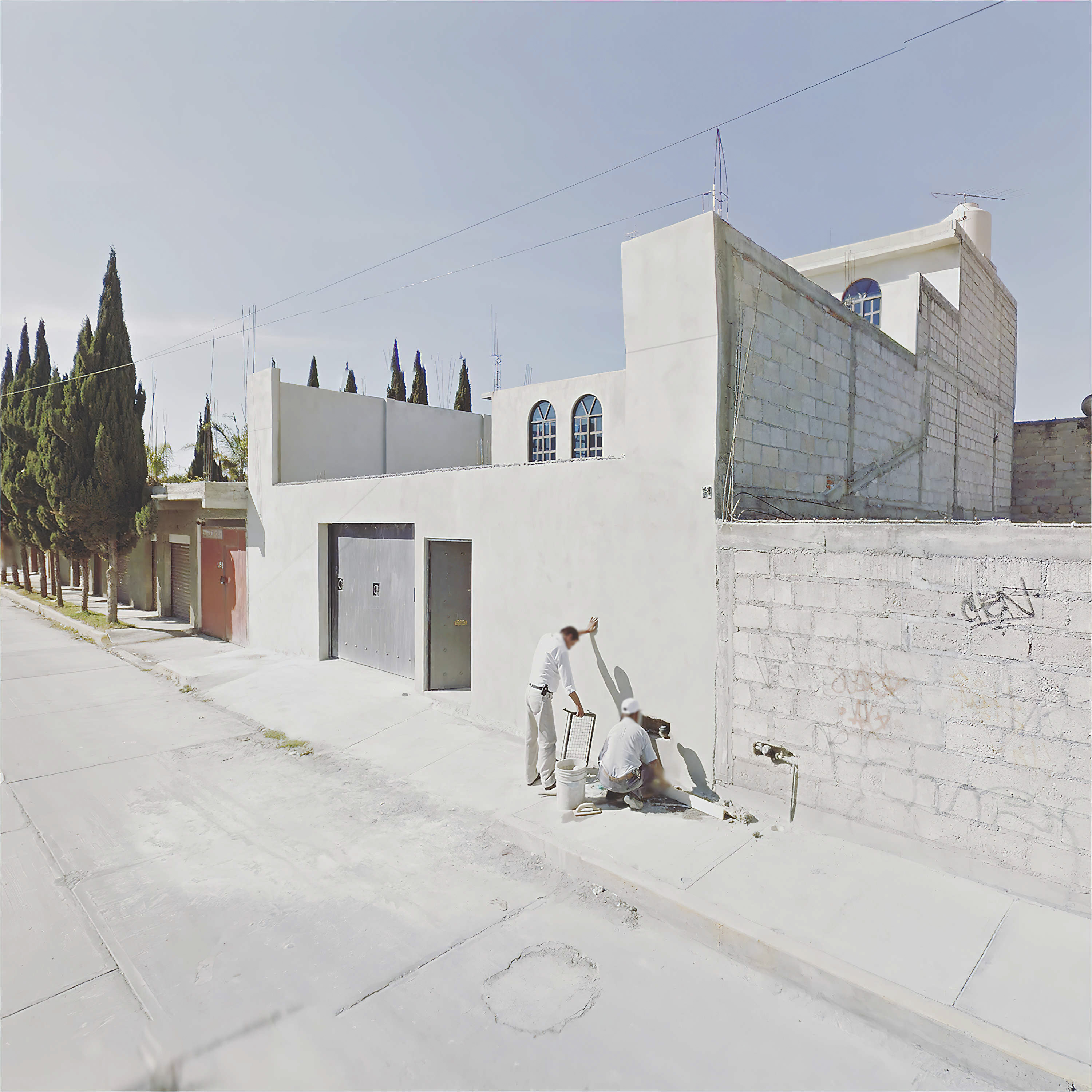 One person kneels on a pavement sidewalk while another person alongside them leads on a white building