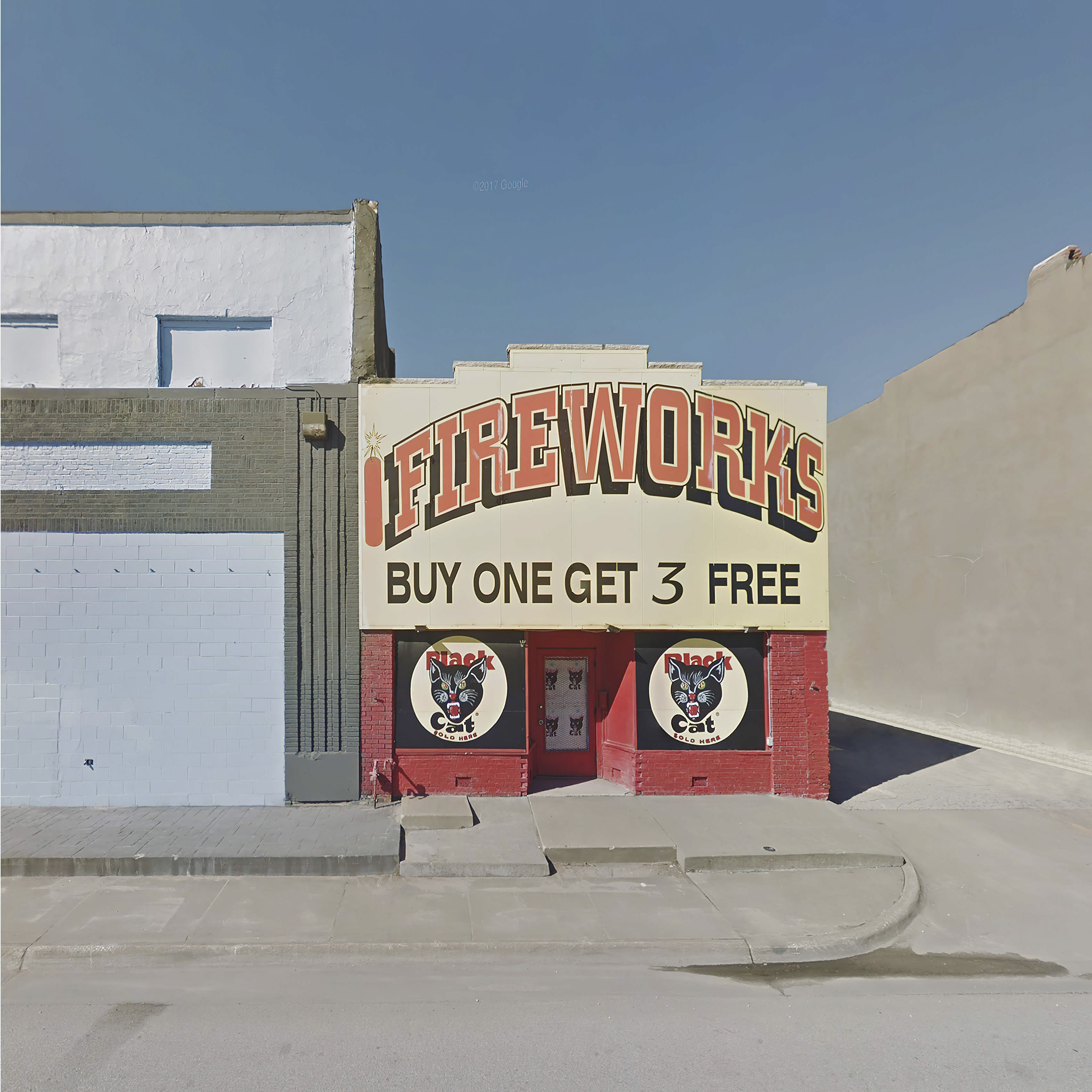 A Black Cat fireworks shop, painted yellow and red, advertising “FIREWORKS buy one get 3 free”, with a blue sky in the background, surrounded by grey pavement sidewalk and walls