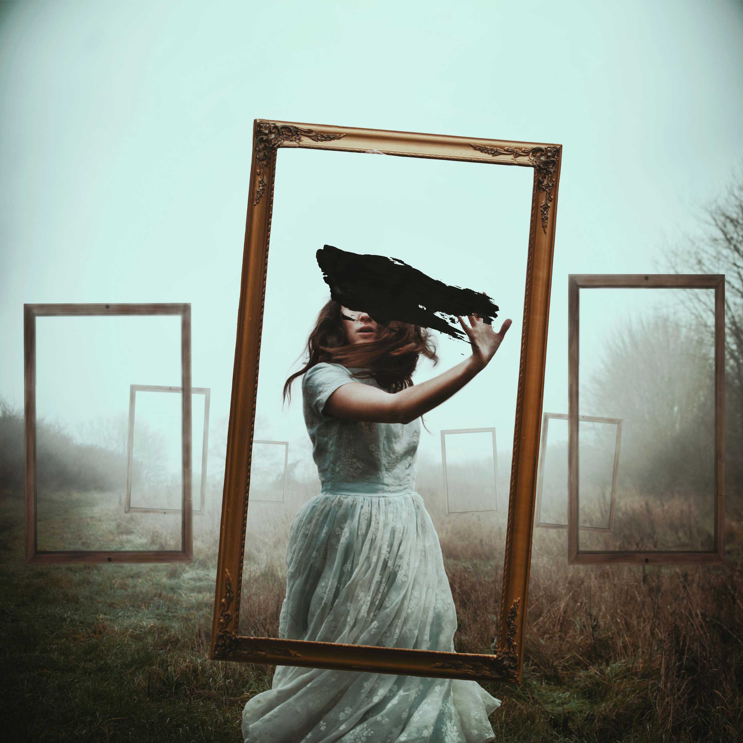 A woman wearing white dress facing the camera smears black paint on a transparent picture frame surrounded by other transparent picture frames in a misty field