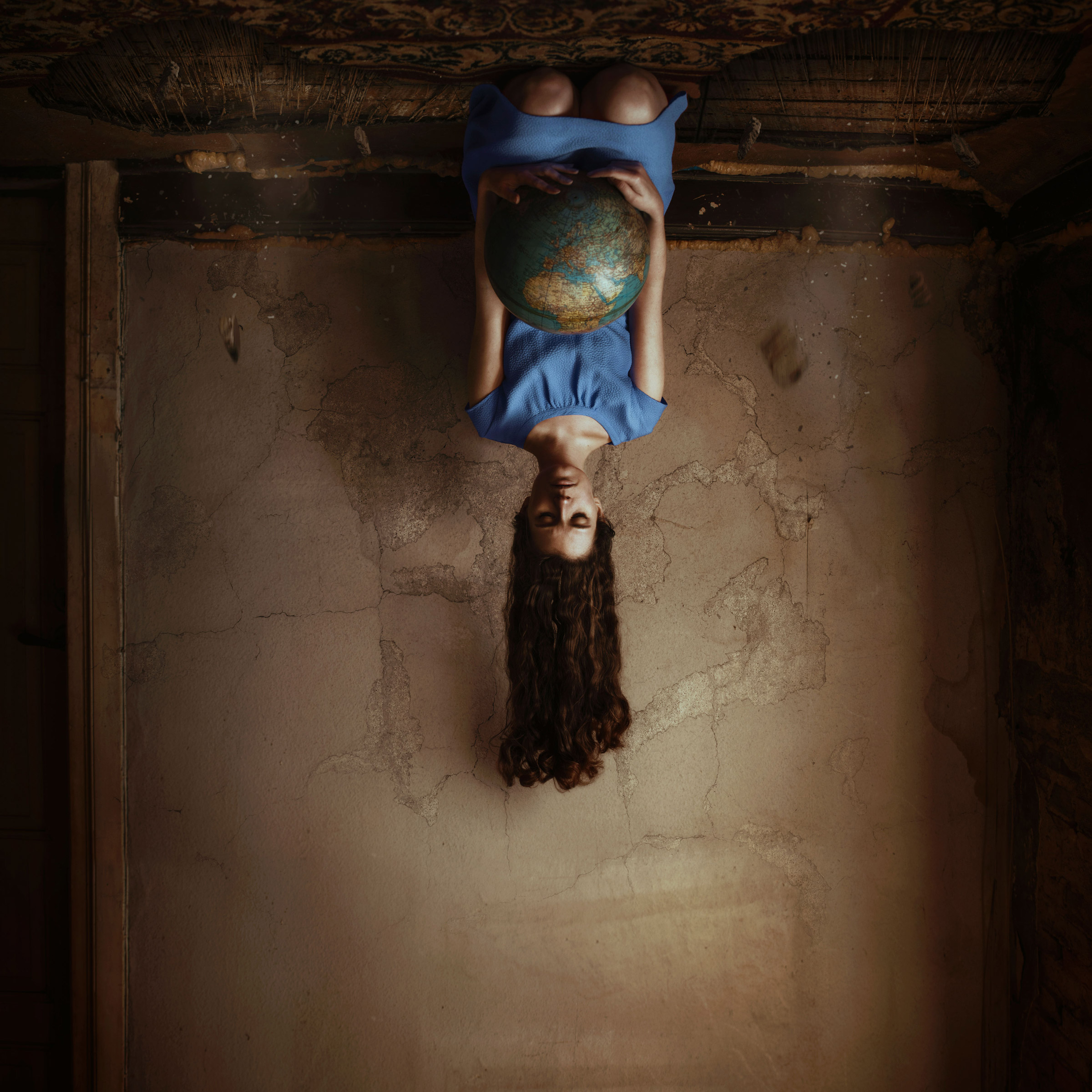A women with long brown hair wearing a blue dress carrying a globe kneels upside down on the ceiling in a brown room
