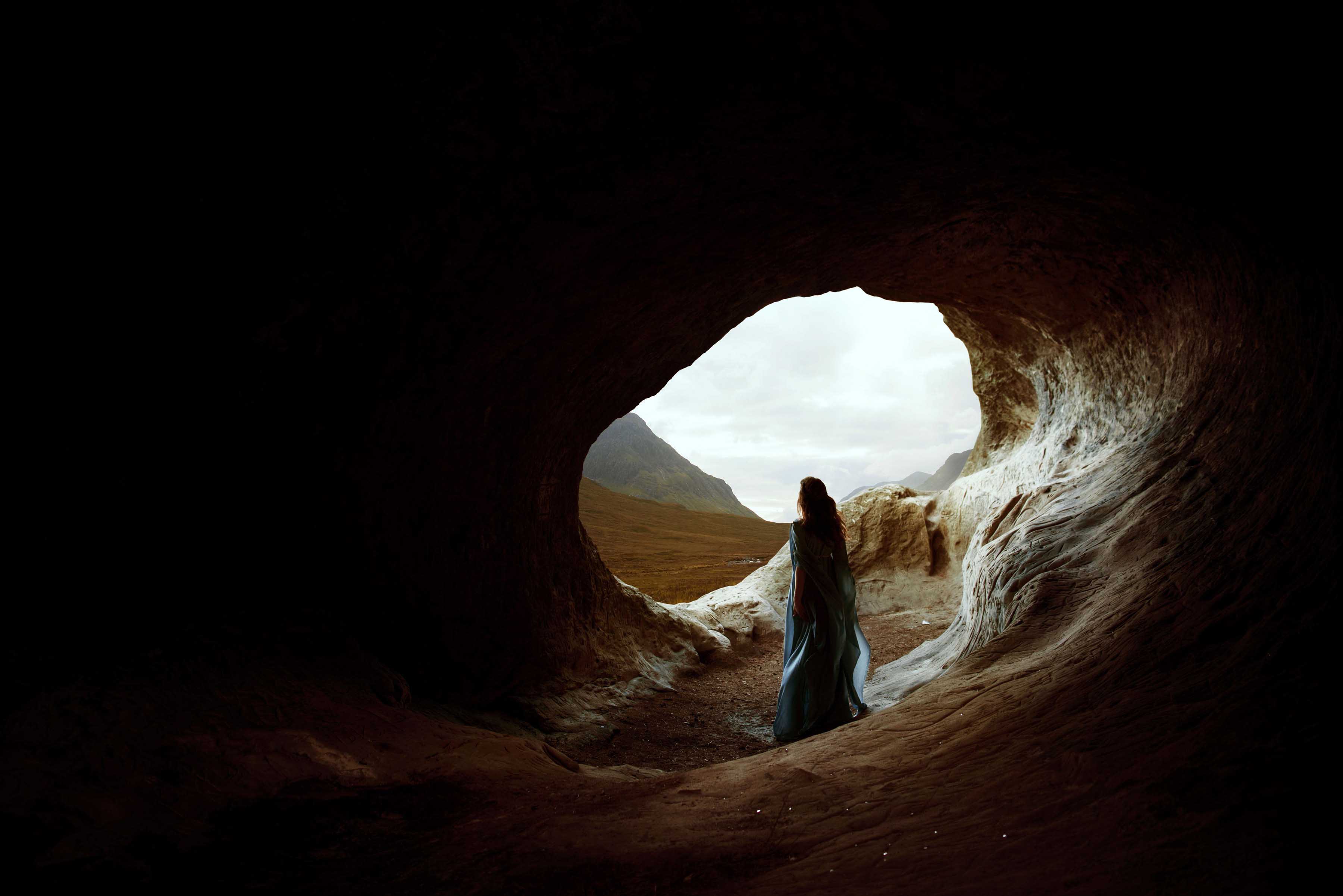 A woman wearing a long blue dress stands inside a dark cave with her back towards the camera, looking out towards the light outside with mountains and sky