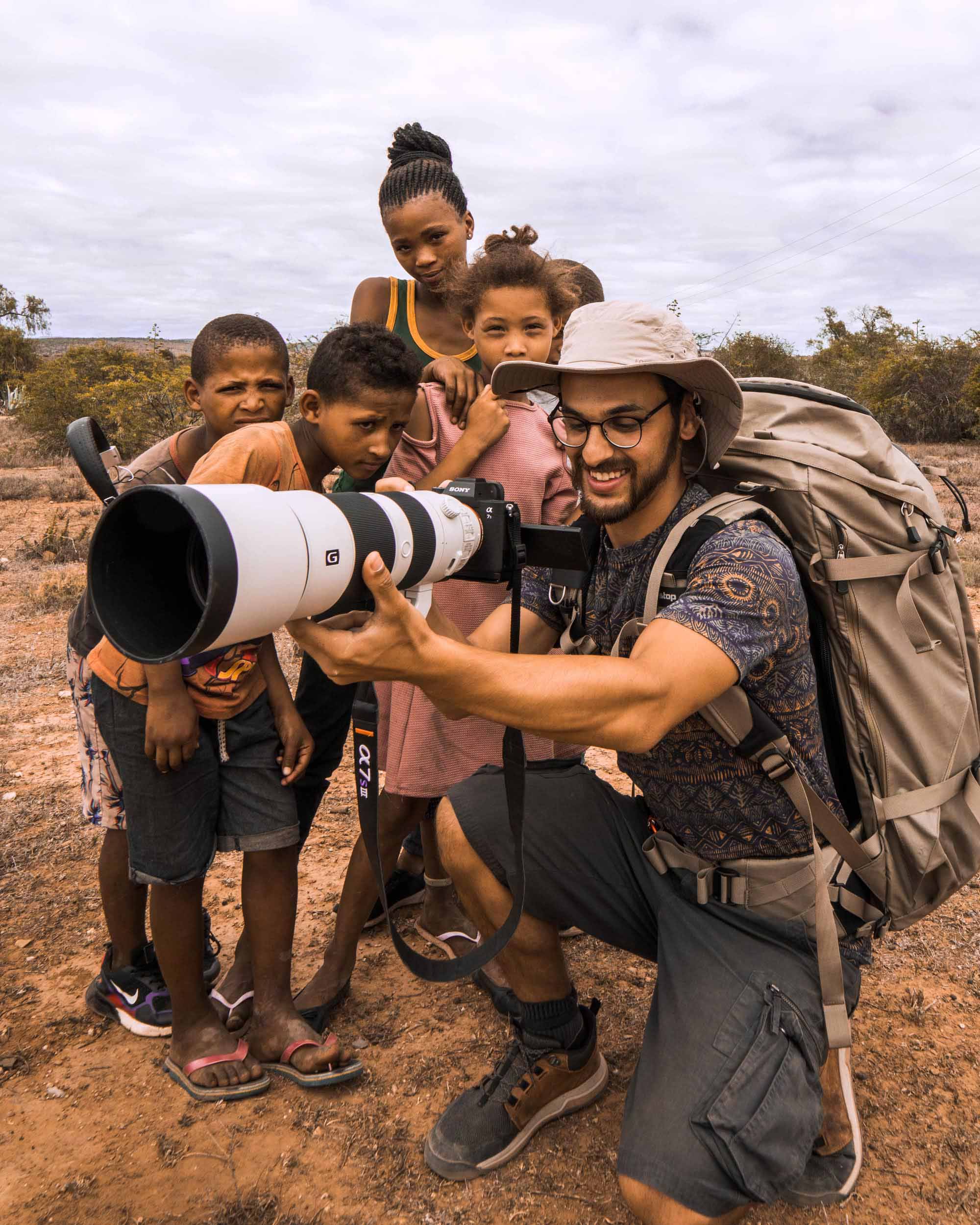 Ein Fotograf mit Hut und Rucksack hält seine Kamera mit einem weißen Teleobjektiv vor einige Kinder