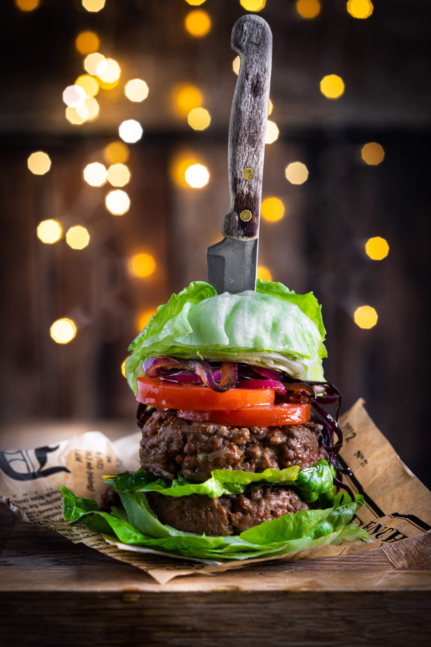 two beef burgers, tomato and onion sitting in between lettuce leaves, being held together with a knife