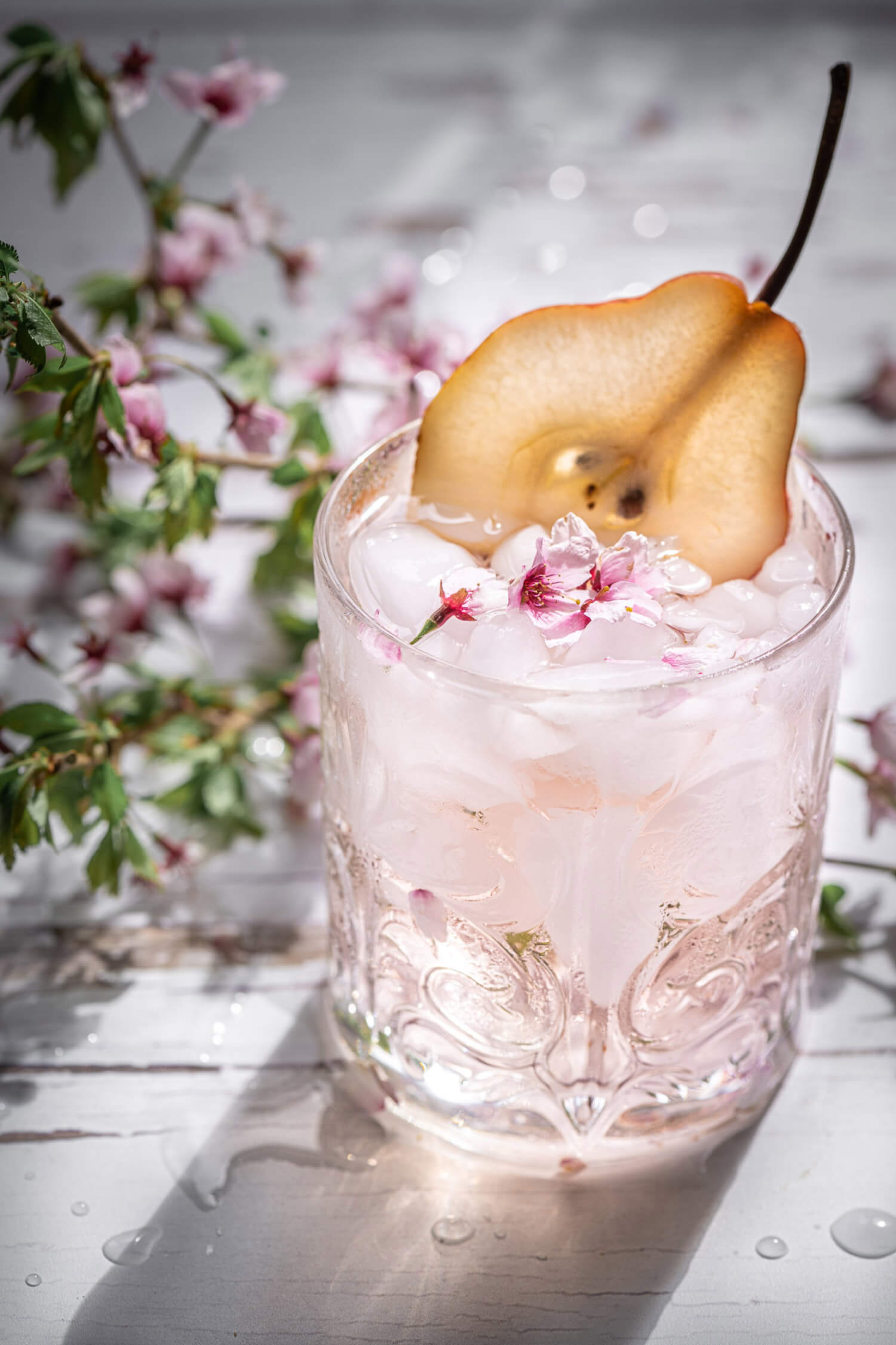 a thinly sliced pear sits in a glass filled with ice, with pink flowers and green leaves