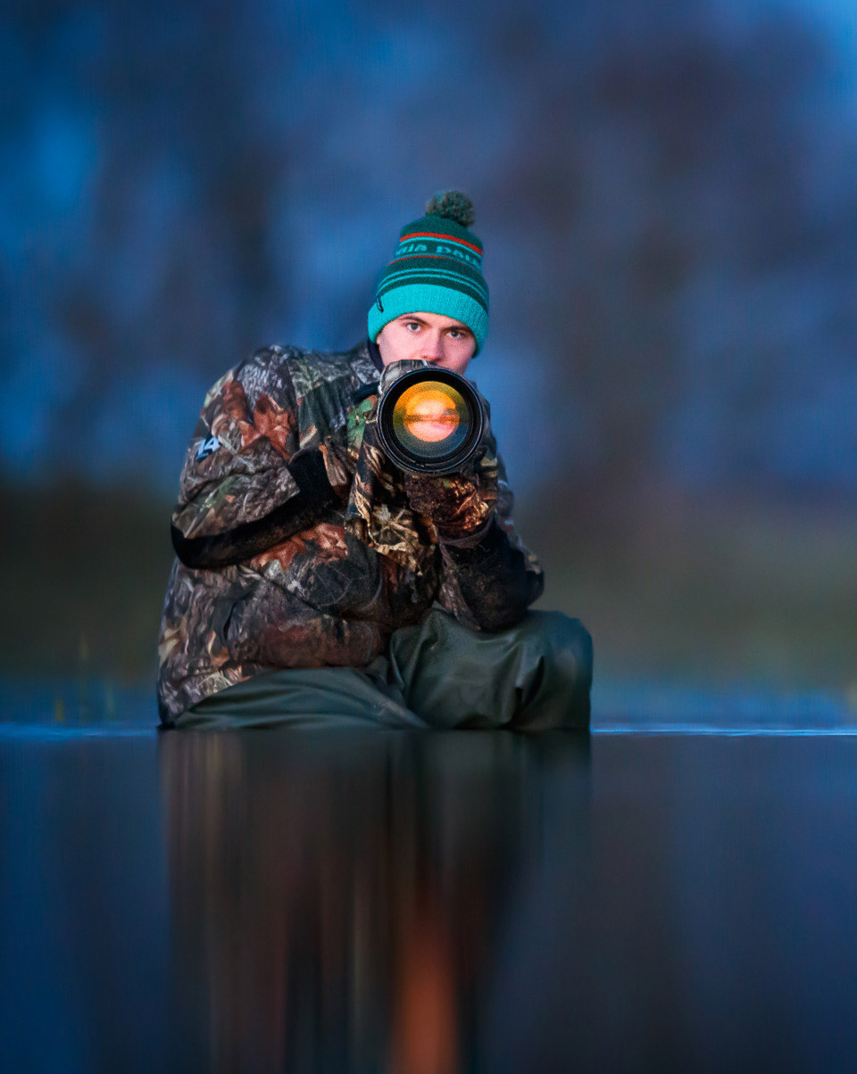 Mateusz Piesiak holding a camera and lens wearing a turquoise hat and camouflage coat while sitting in a river