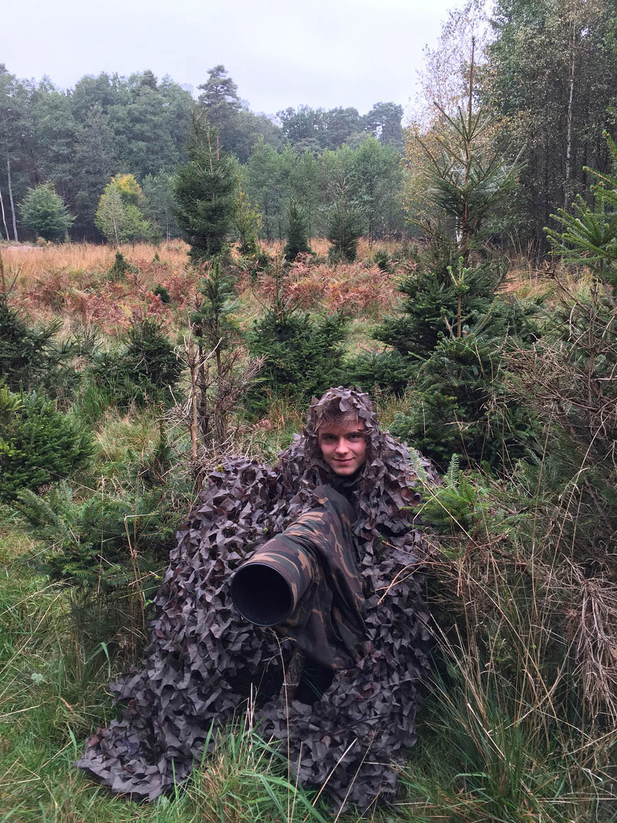 Wildlife fotograaf Mateusz Piesiak in het bos in camouflagekleding met een lens in de hand
