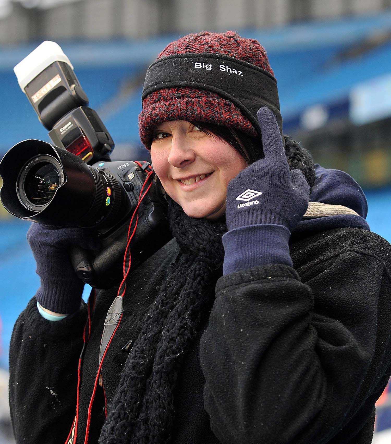 Sharon Latham holds a camera whilst pointing at her hat that has Big Shaz written on it.