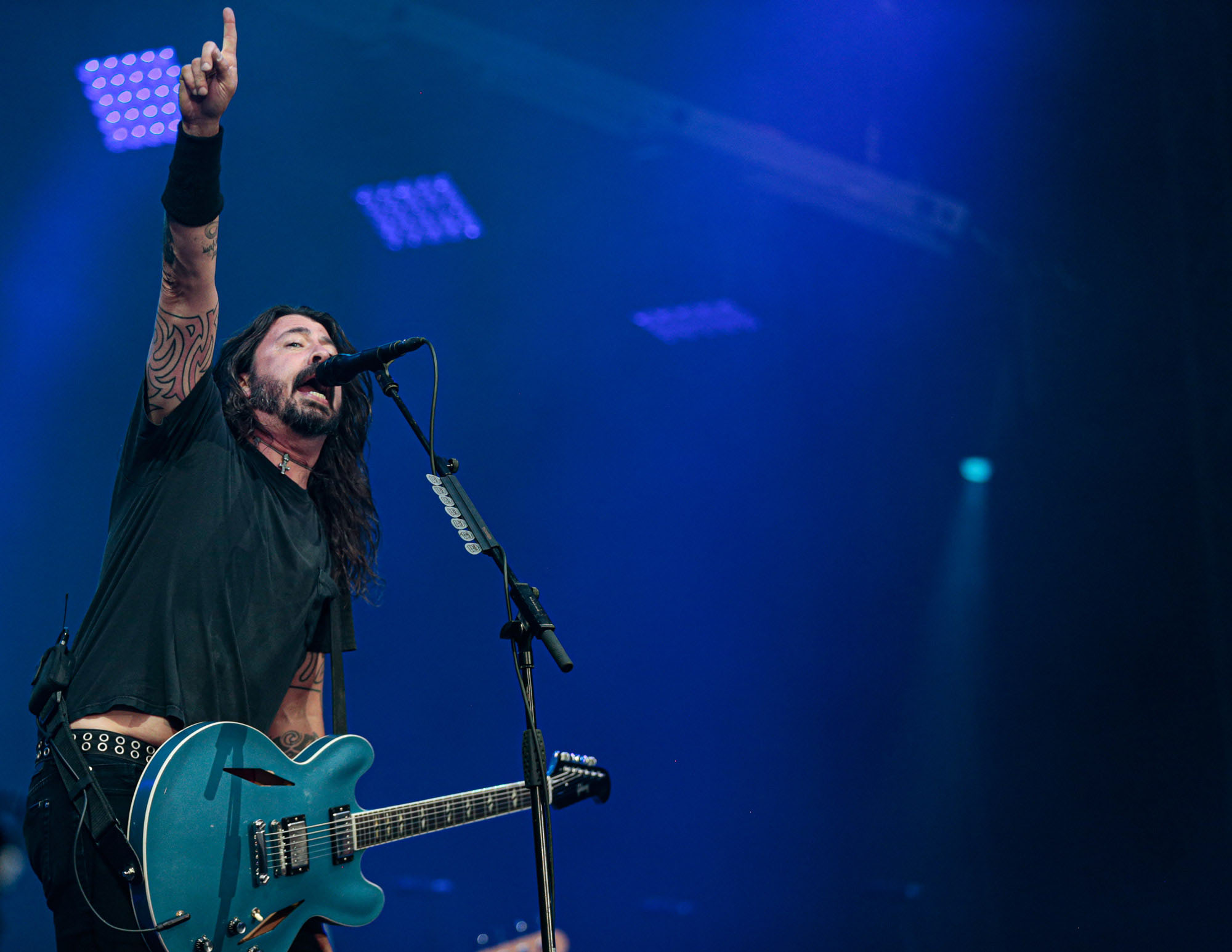Dave Grohl sings into a microphone on stage, he is holding a blue guitar and has one arm raised, pointing in the air.