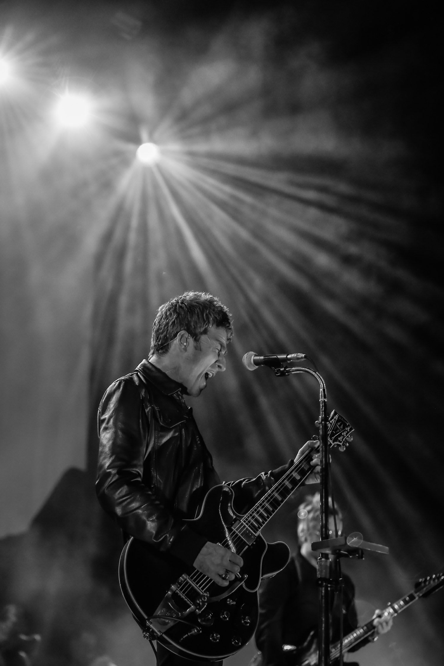 A black and white image of Noel Gallagher playing the guitar whilst singing.
