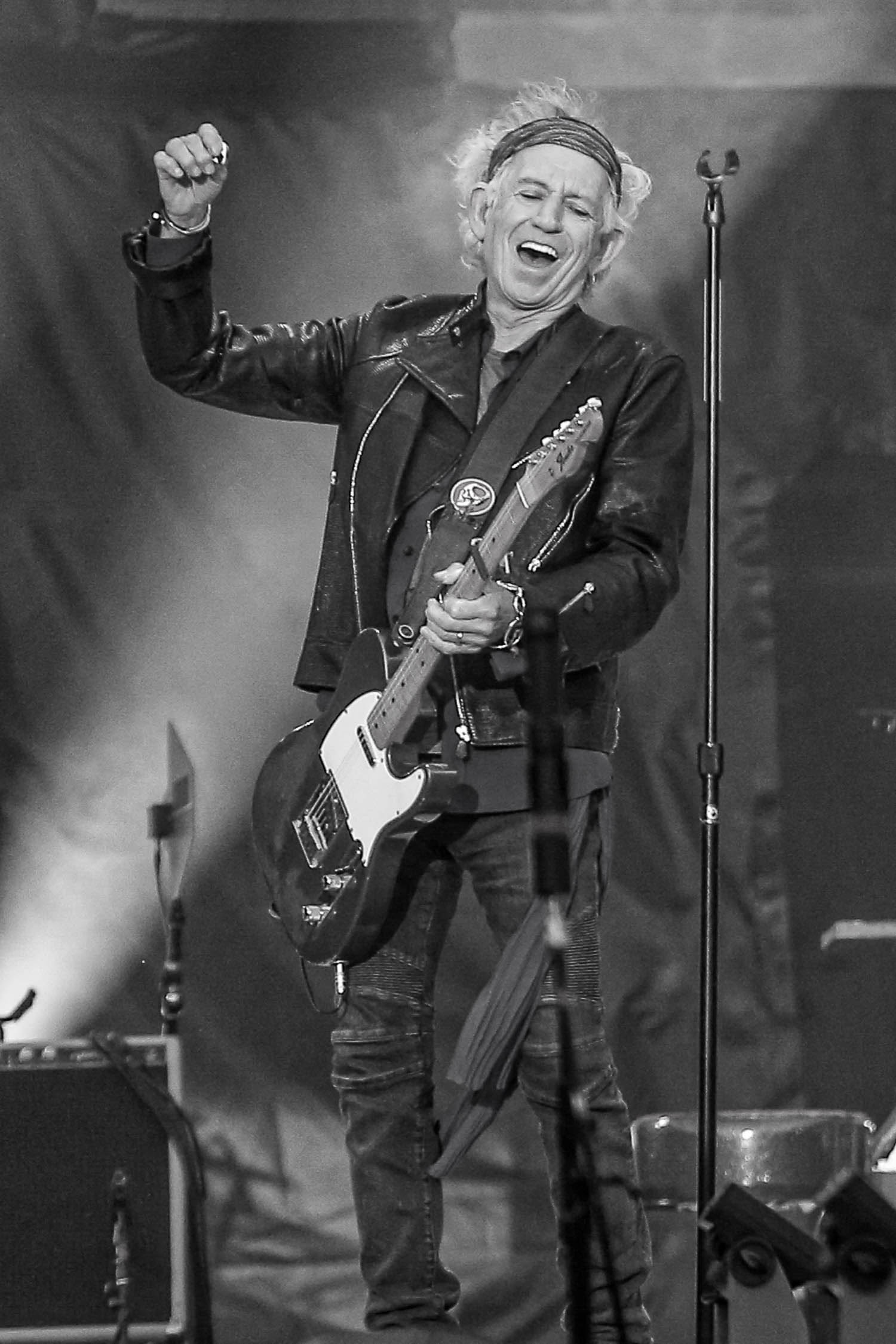 A black and white image of Keith Richards smiling as he plays the guitar live on stage.