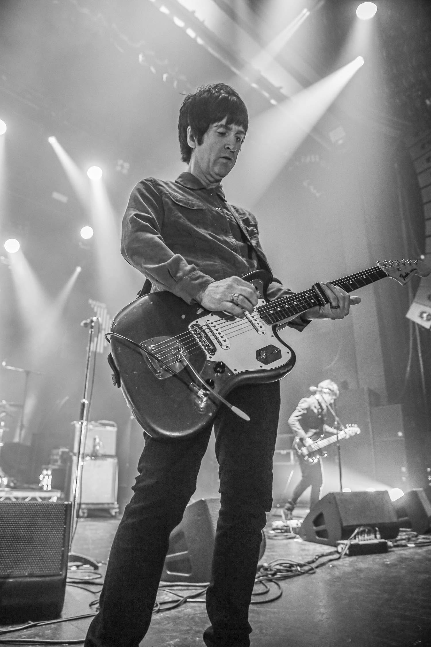 A black-and-white image of Johnny Marr, playing guitar on stage. Speaker, microphones and another guitarist are in the background.