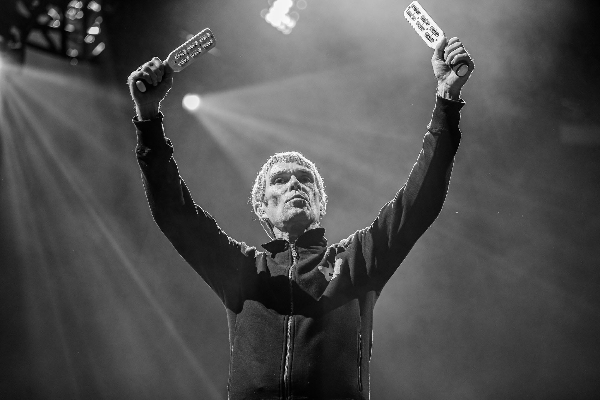 A black and white image of Ian Brown with his arms raised above his head, with a jingle stick in each hand.