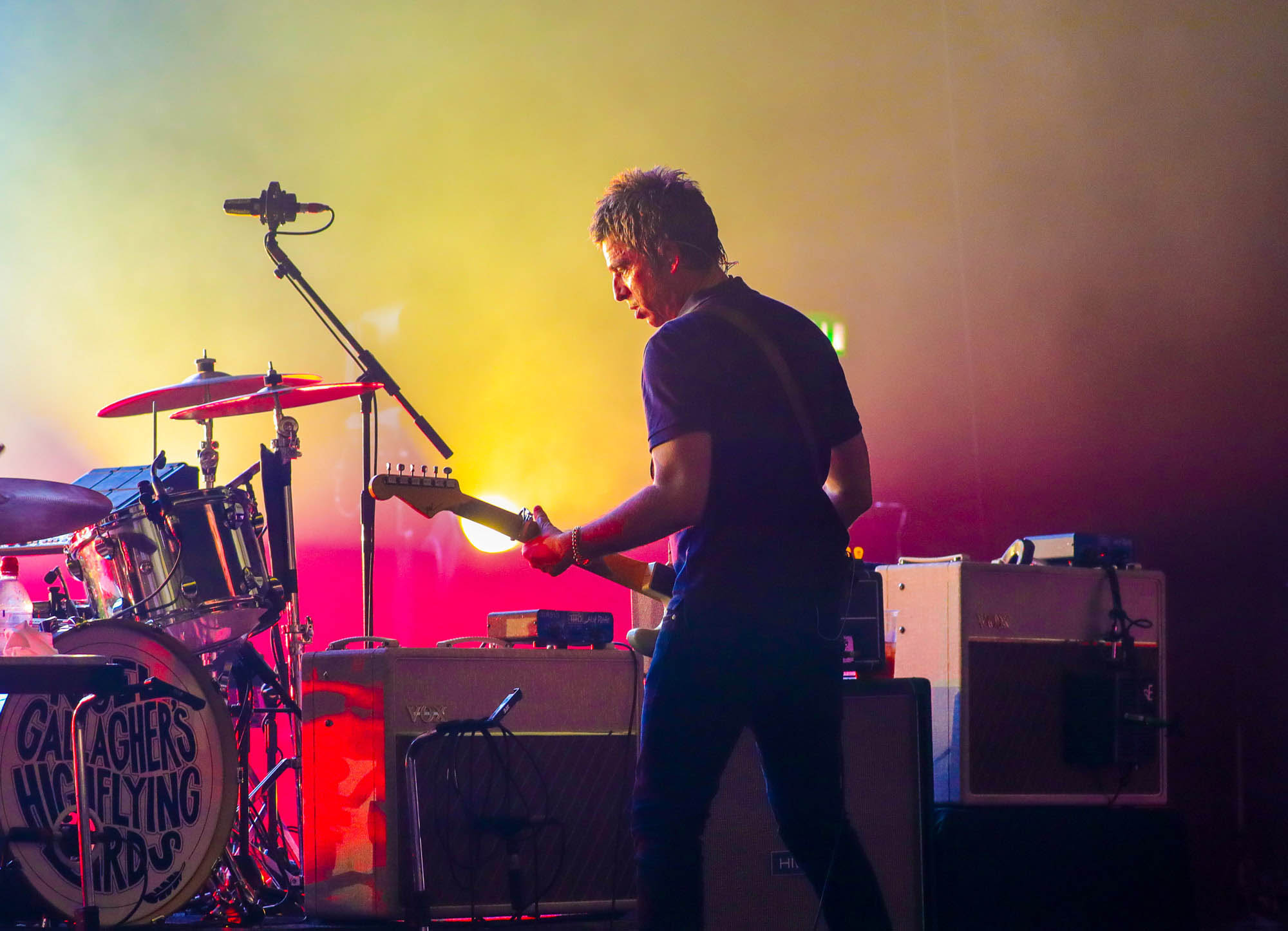 Noel Gallagher playing a guitar stands with his back to camera while performing live on stage with a drum kit in the background.
