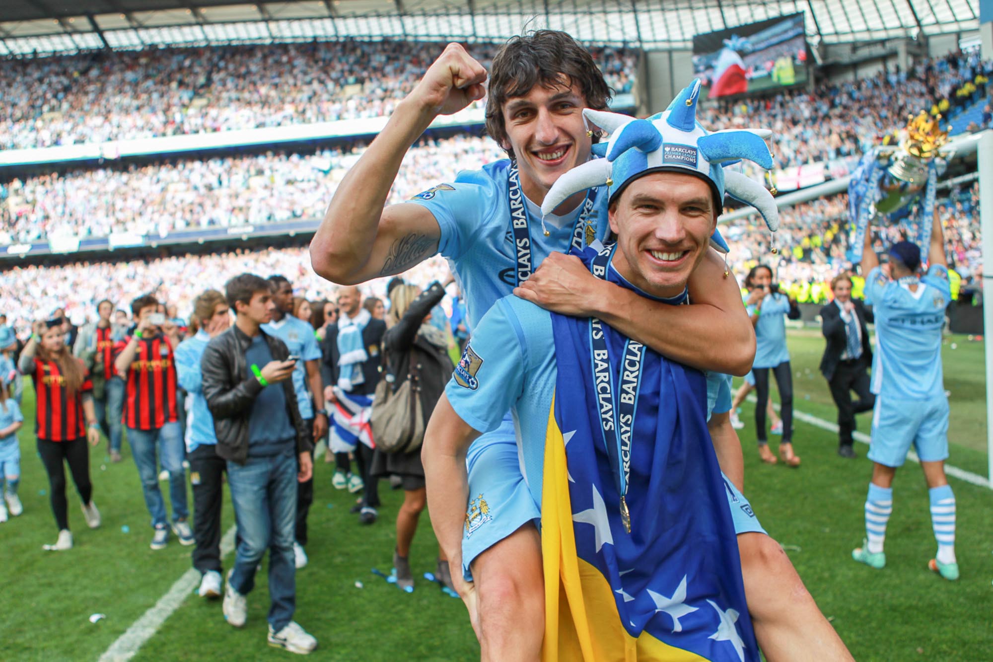 A smiling Edin Dzeko carries Stefan Savić on his back, with players celebrating in the background.