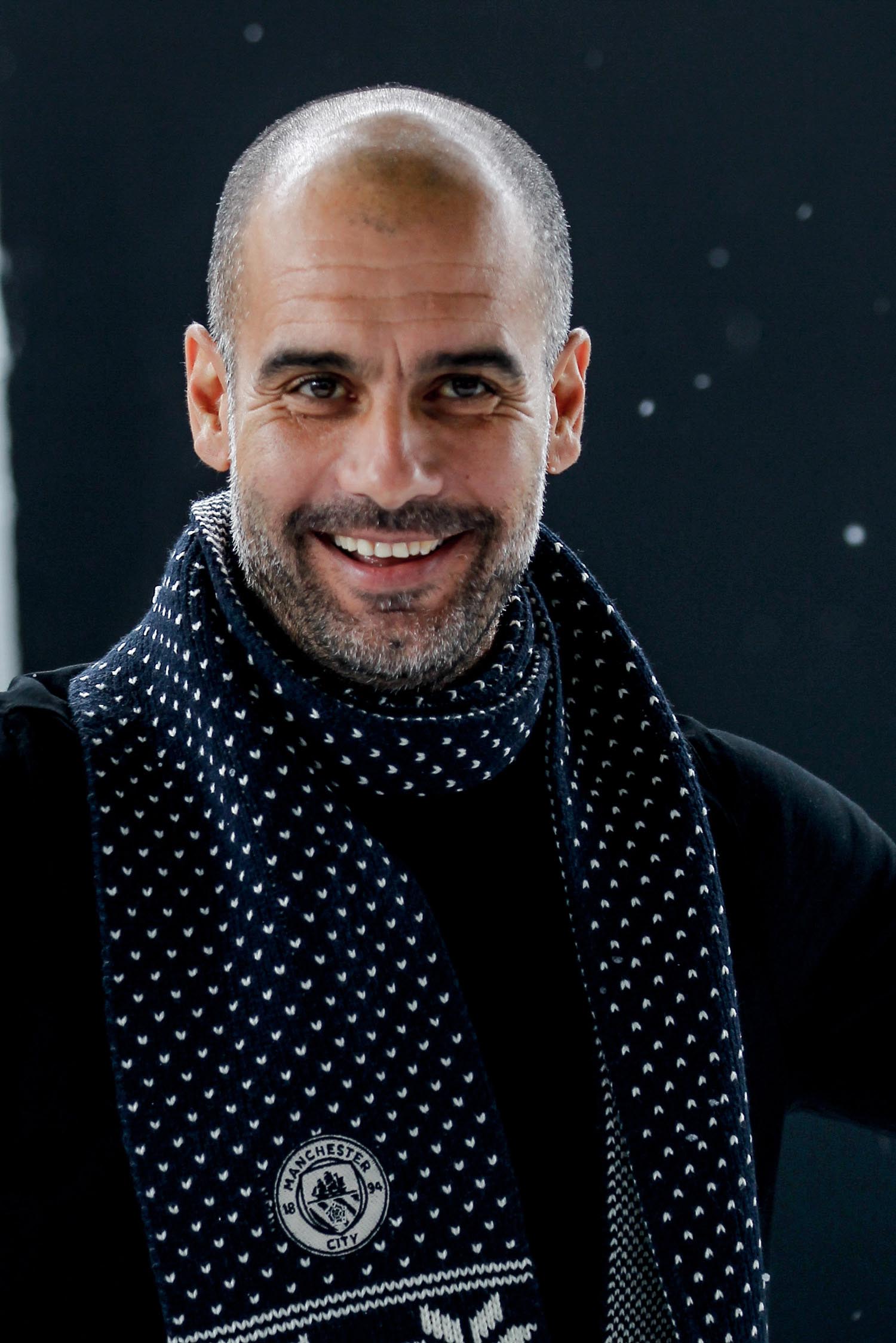 Pep Guardiola smiling as he wears a black and white Manchester City scarf