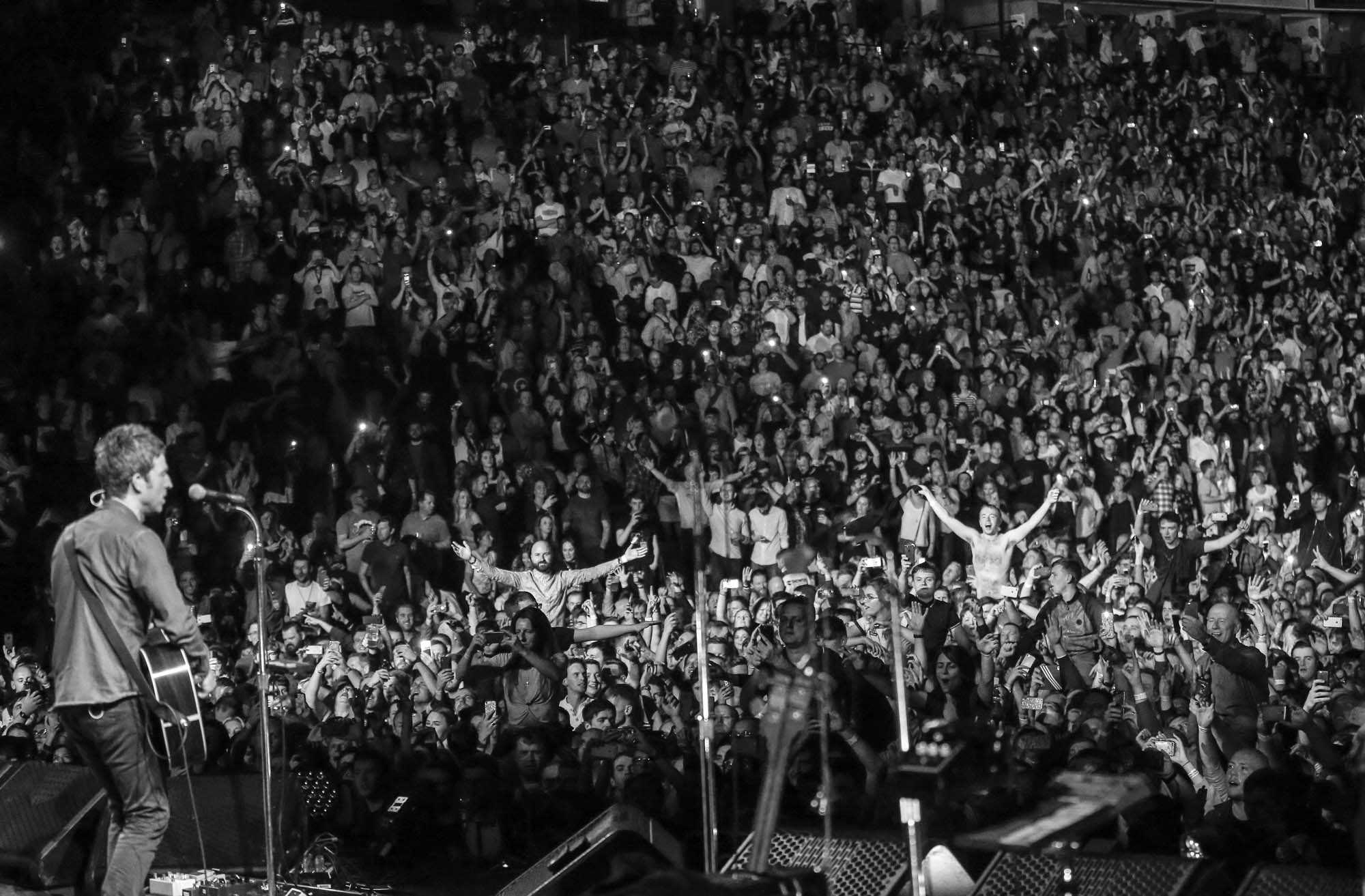 A black and white image of Noel Gallagher on stage singing, with a large crowd in the background.